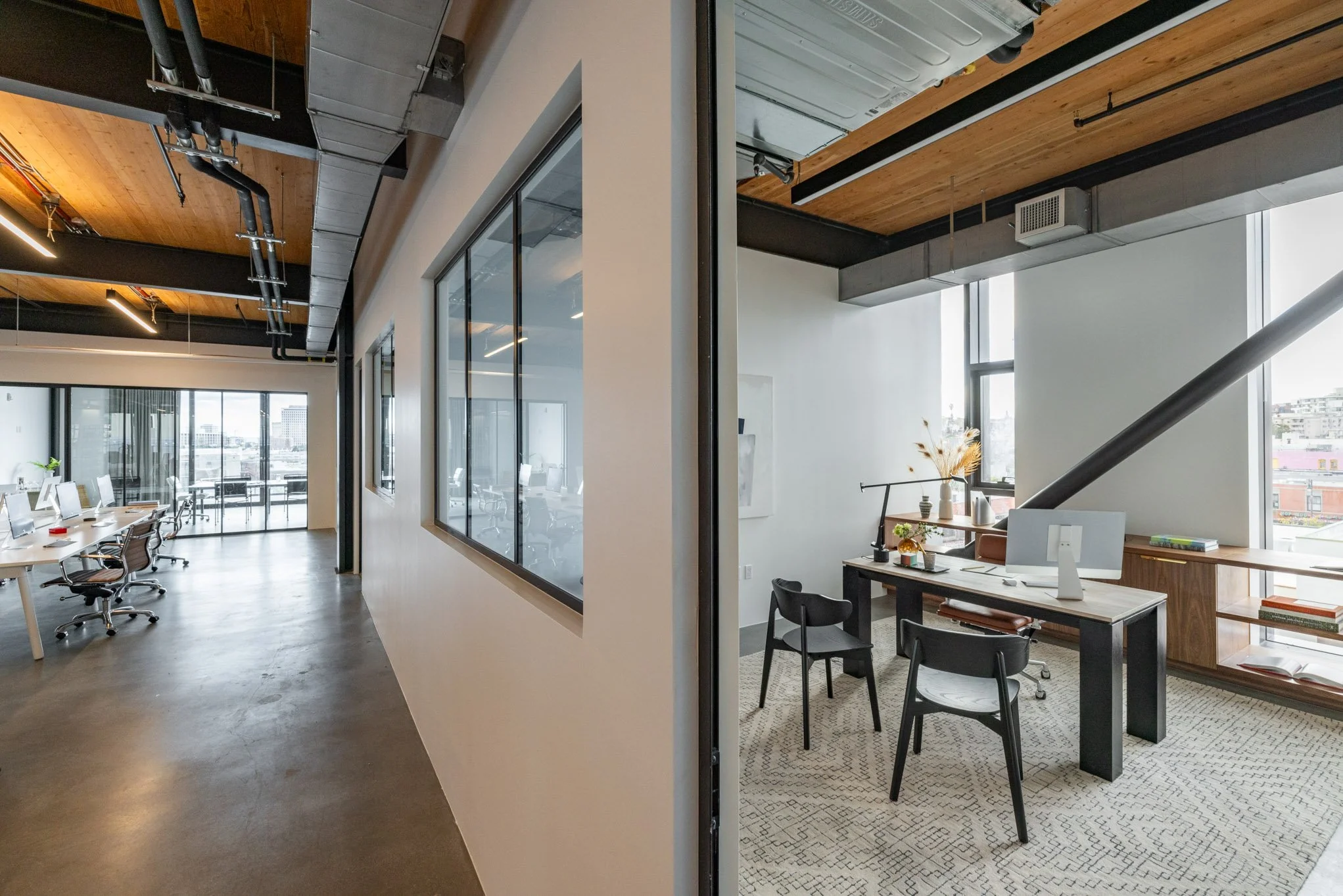 Split view of modern office space with open work area containing desks and chairs on the left, and a private office with a desk, chairs, and large windows on the right.