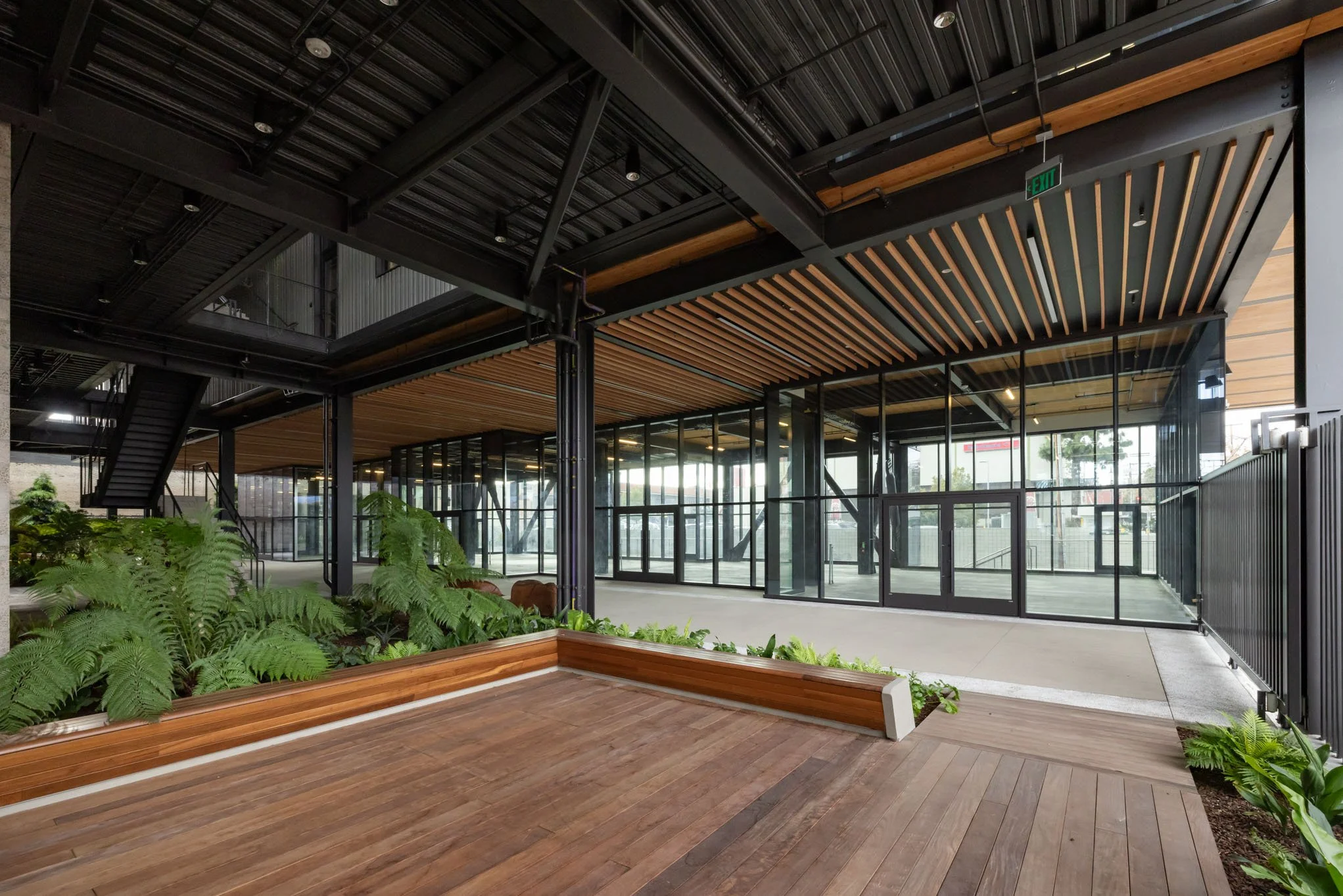Modern indoor space with glass walls, wooden ceiling, wooden and concrete flooring, greenery, and metal staircase.