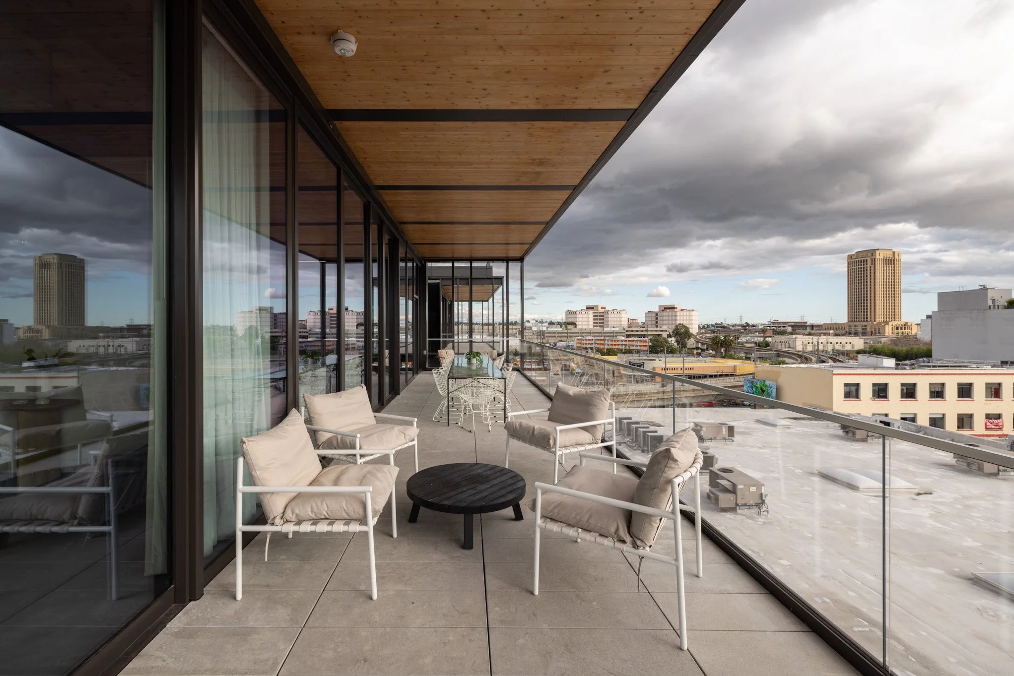 A spacious balcony with beige chairs and a black coffee table, overlooking an urban cityscape with tall buildings, under a cloudy sky.