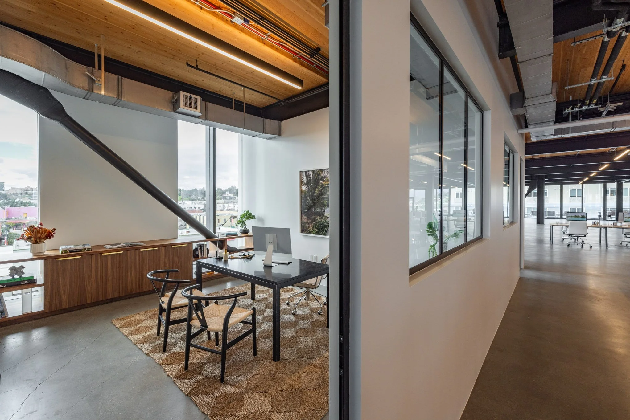 Modern office space with large windows, a desk with a desktop computer, two black chairs, bookshelf, plants, and a rug. The office has a contemporary design with wood, concrete, and glass elements.