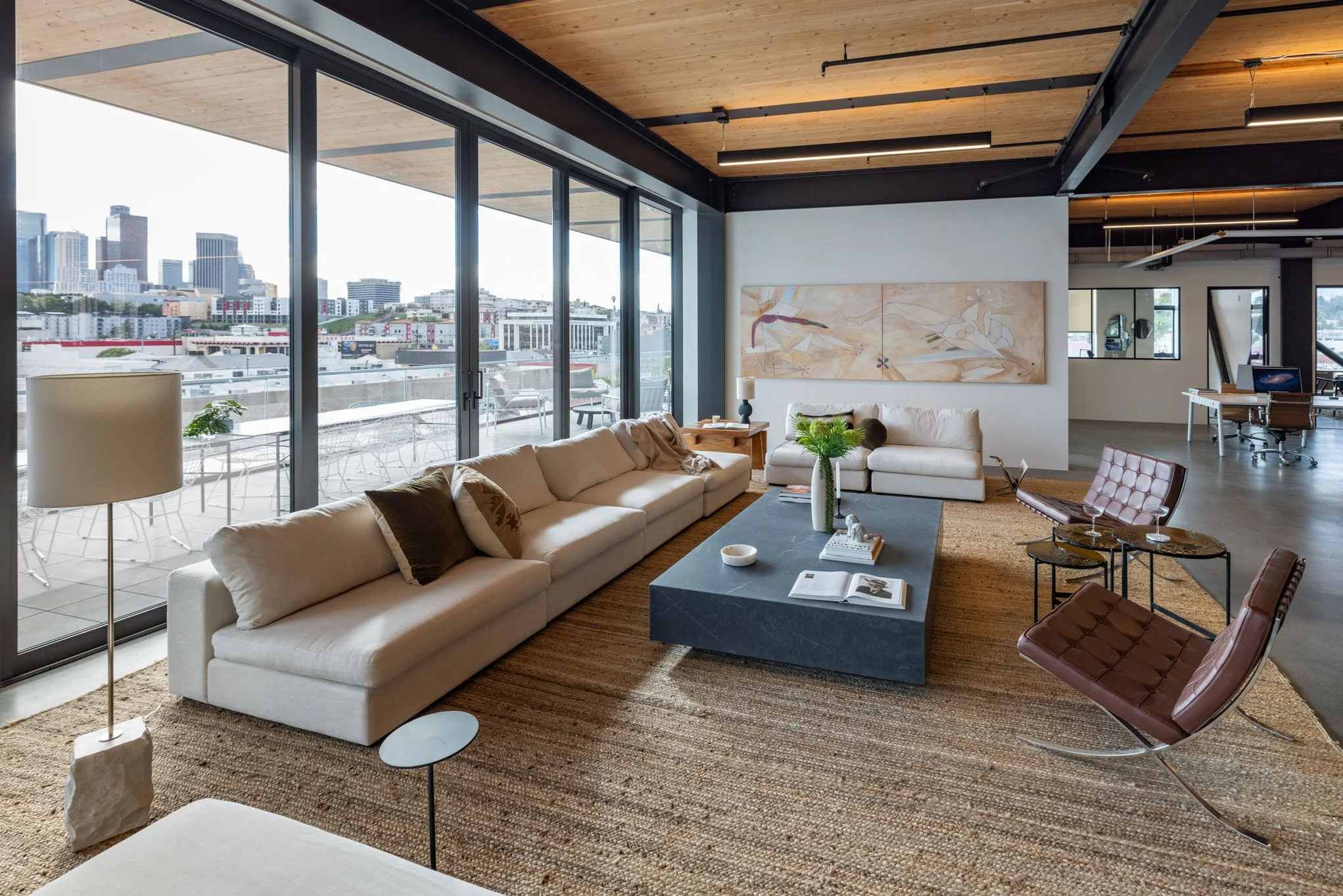 Modern living room with beige sectional sofa, leather lounge chair, large black coffee table, area rug, large windows with city view, abstract wall art, and decorative plants.