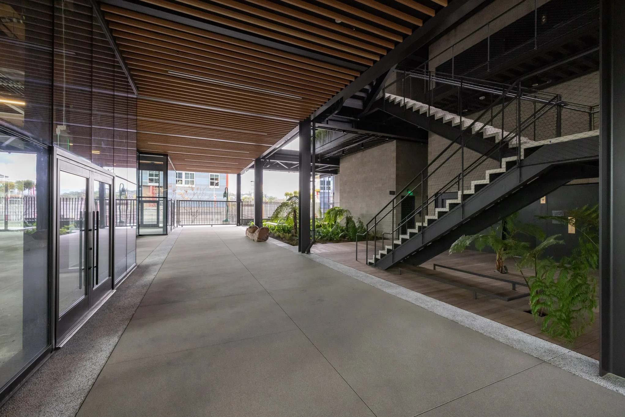Modern indoor lobby area with glass doors, concrete flooring, staircase with black metal railing, wooden ceiling, and indoor plants.