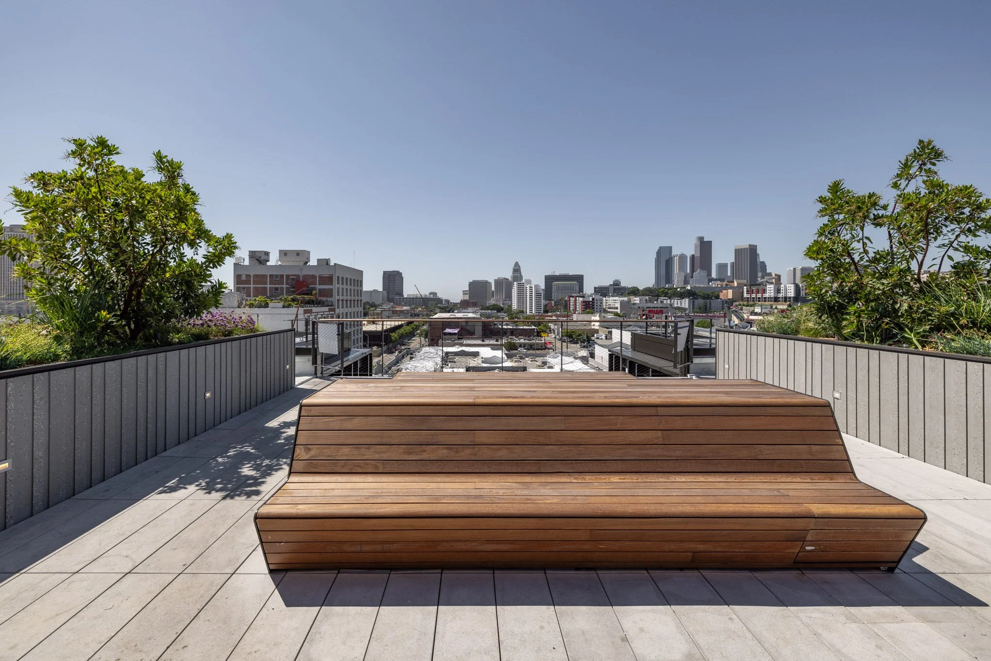 Empty rooftop deck with a wooden bench and city skyline in the distance under a clear blue sky.