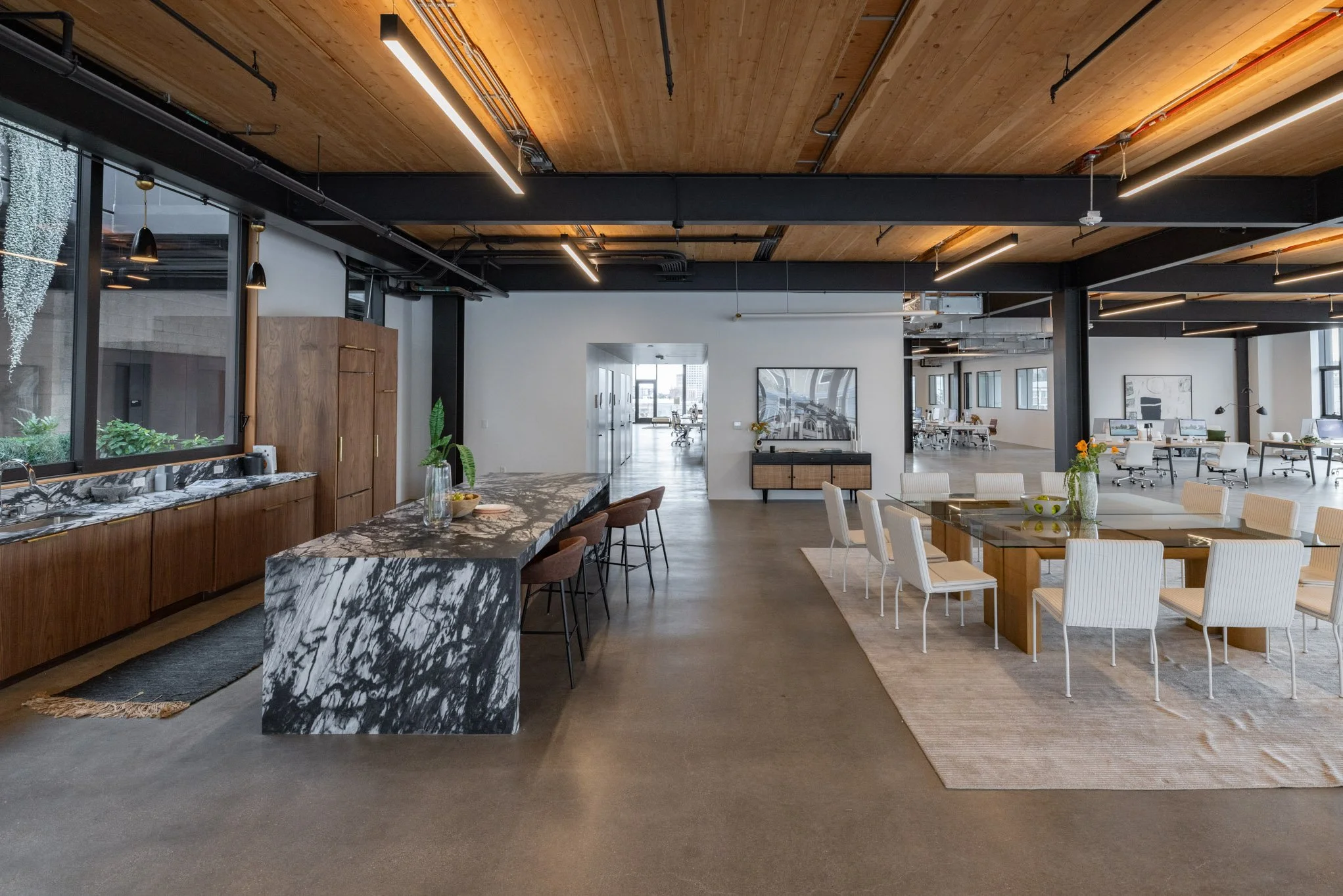 Modern open-plan office space with a kitchen area featuring a marble countertop and cabinets, a large dining table with white chairs, and multiple workstations with desks and computers in the background.