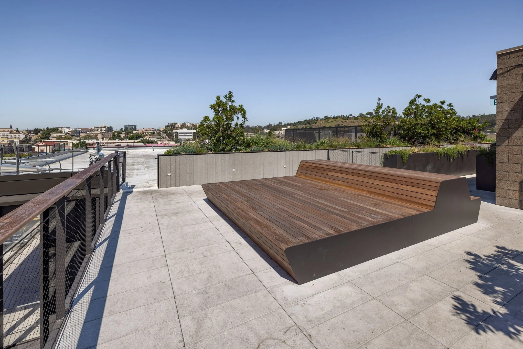 Rooftop balcony with wooden seating, black metal railing, and a view of city buildings and greenery under a clear blue sky.