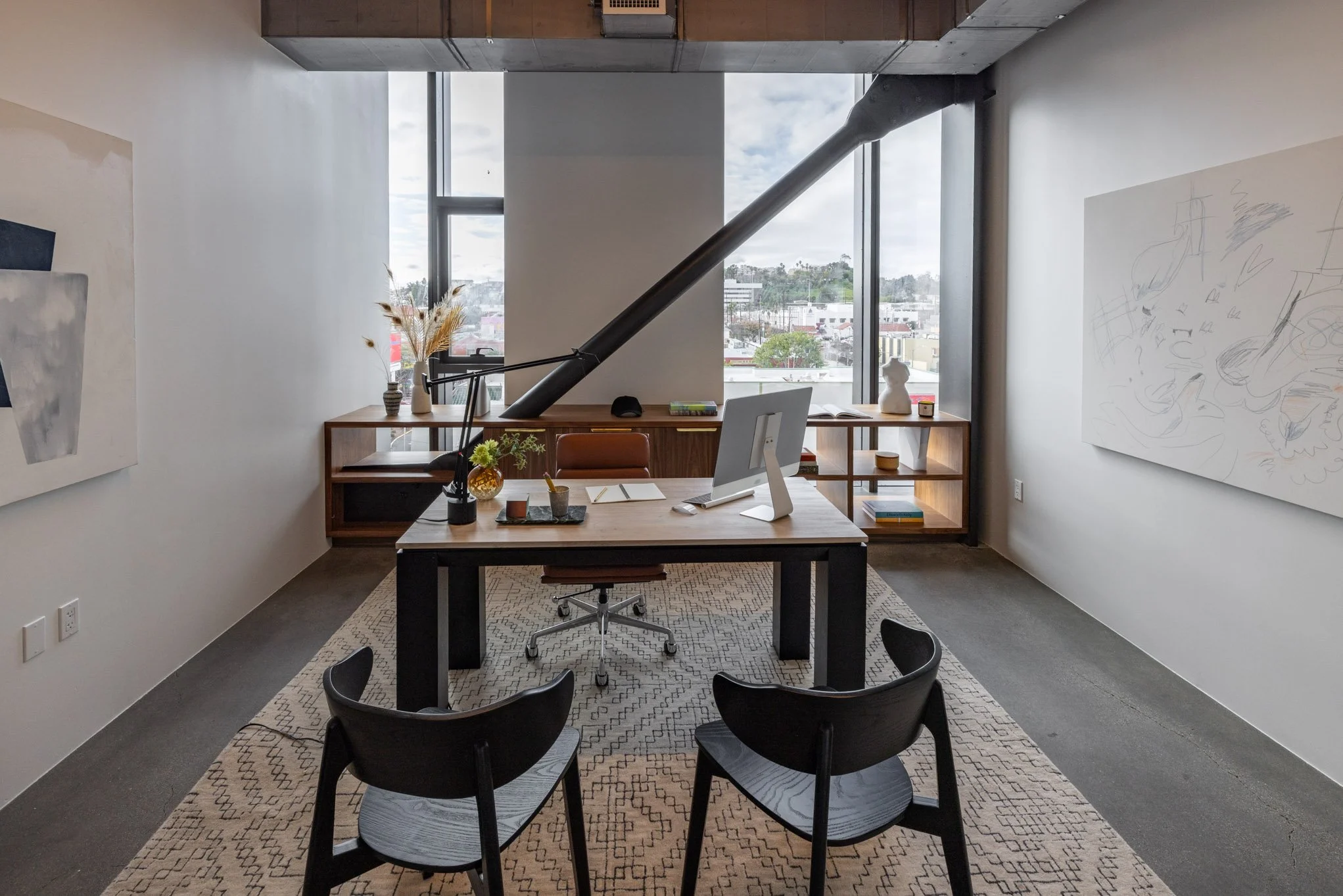 Modern office with a wooden desk, black chairs, and a large window overlooking a cityscape. Decor includes vases with dried flowers, books, and artwork on the walls.