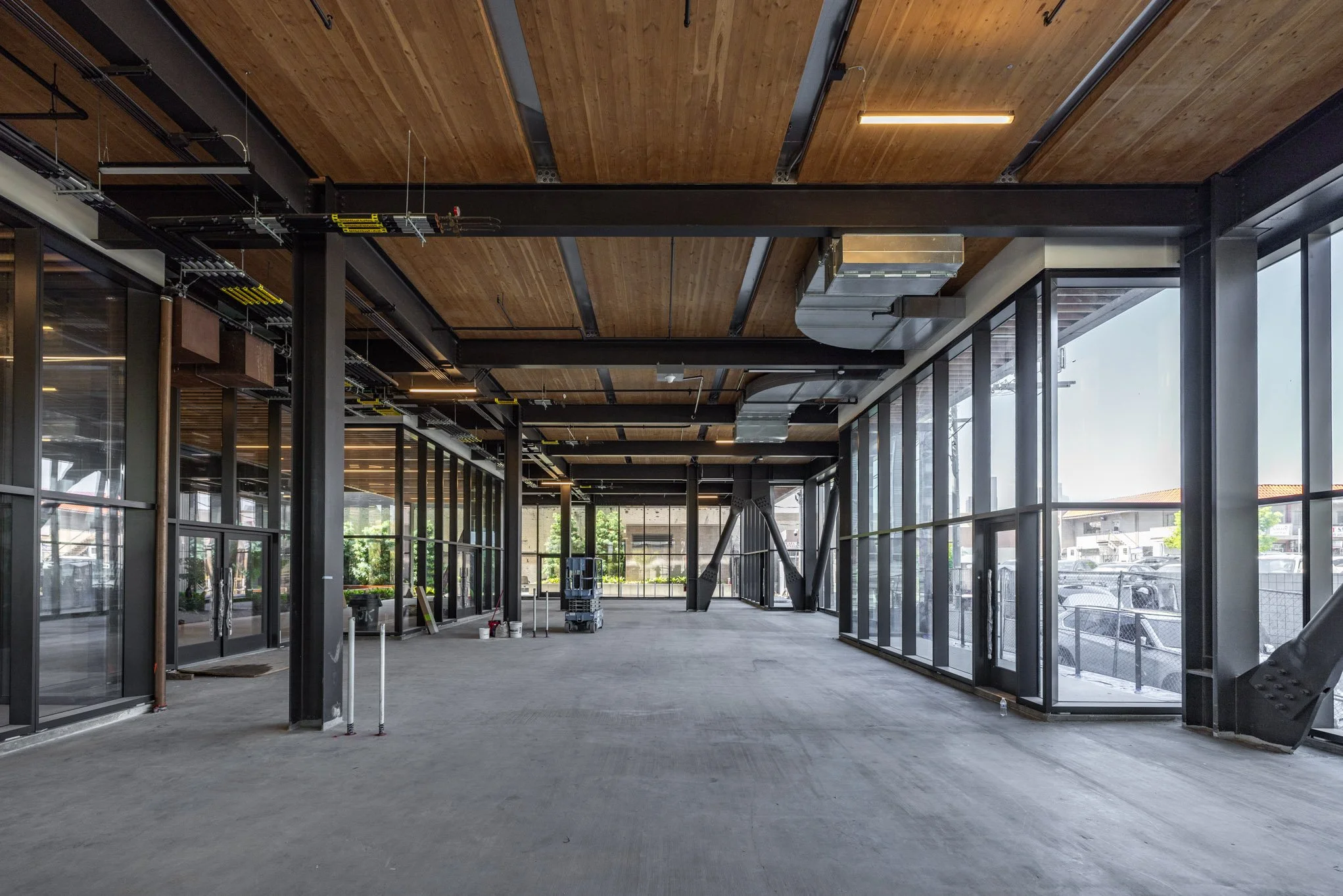 Interior of a commercial building under construction with large glass windows, exposed ceiling with wood and metal, and construction equipment inside.