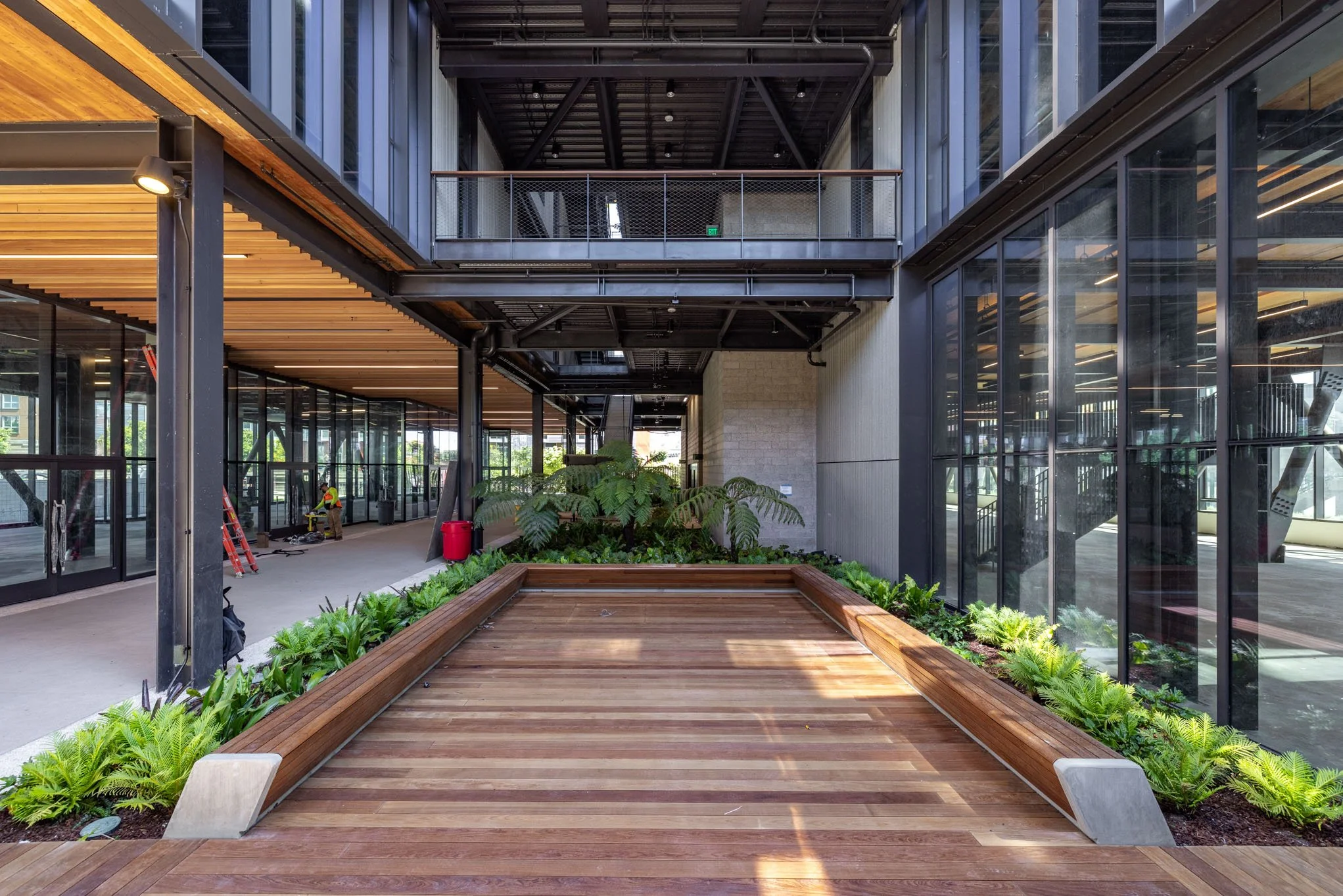 Indoor courtyard with wooden walkway, lush green plants, and glass walls in a modern building.