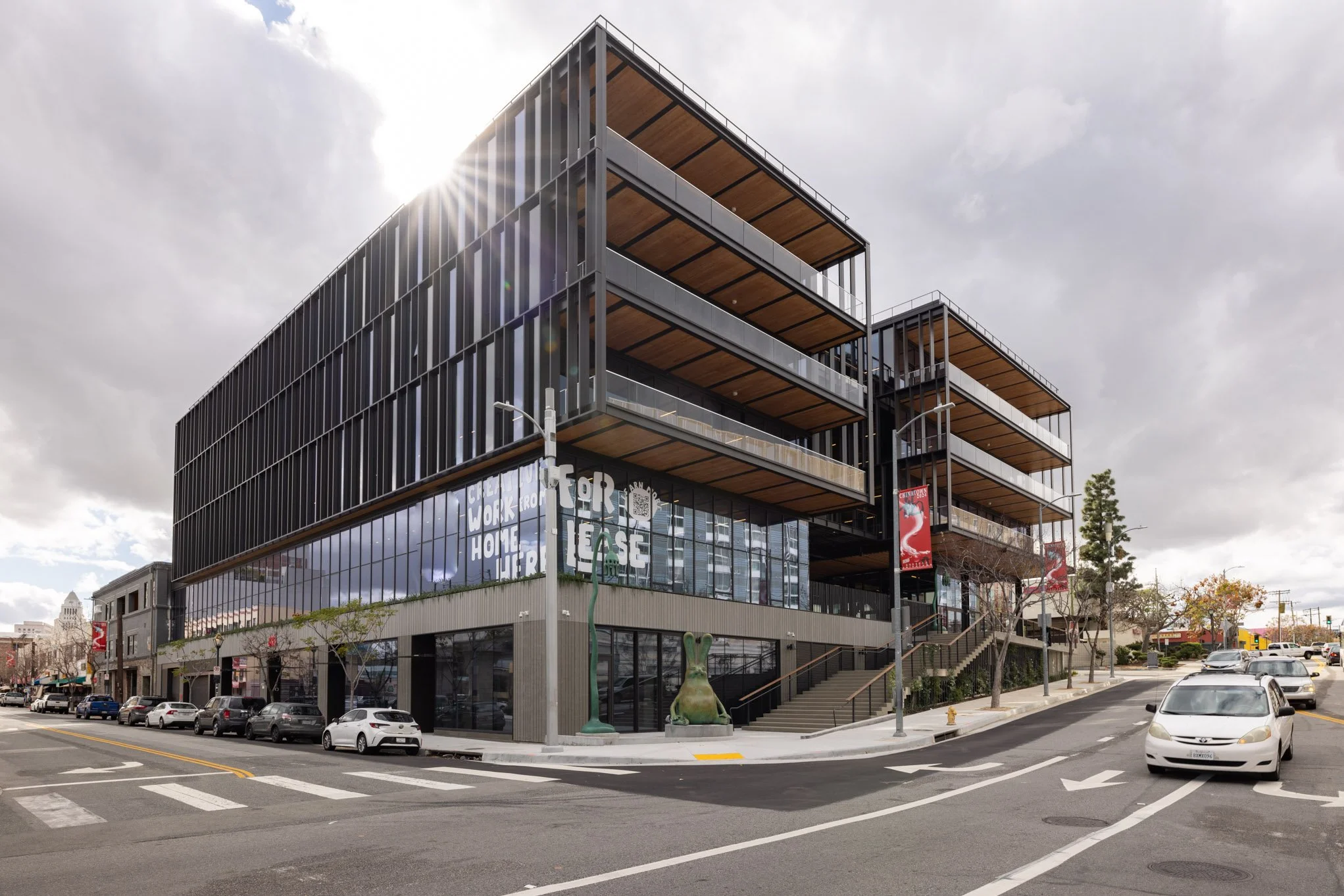 Modern multi-story commercial building with large glass windows and black exterior on a city street, with parked cars and trees, overcast sky.