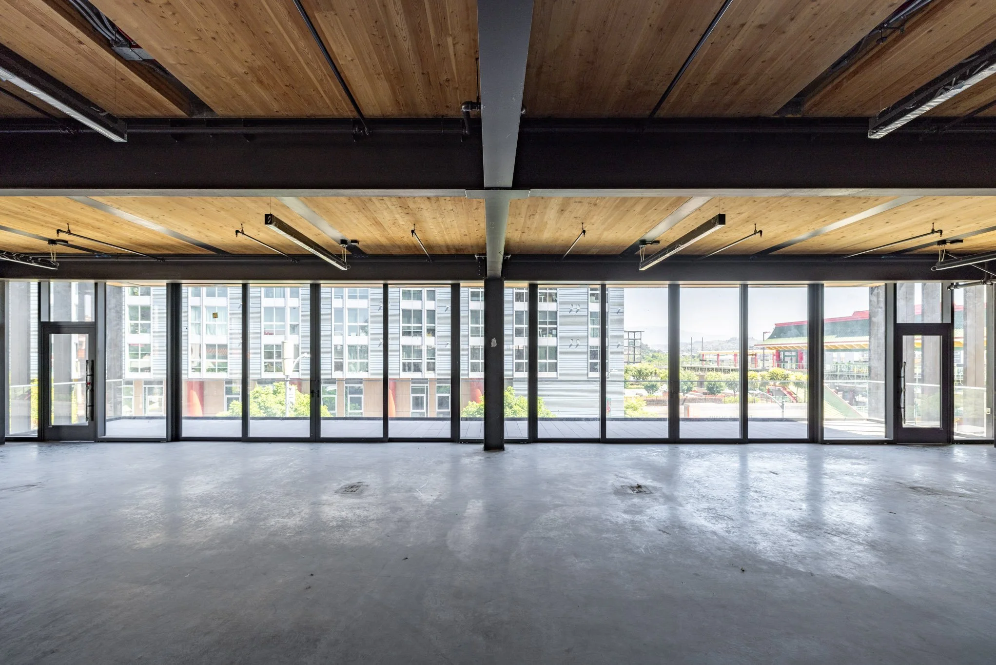Empty commercial space with large floor-to-ceiling windows, wooden ceiling panels, and unfinished concrete floor.