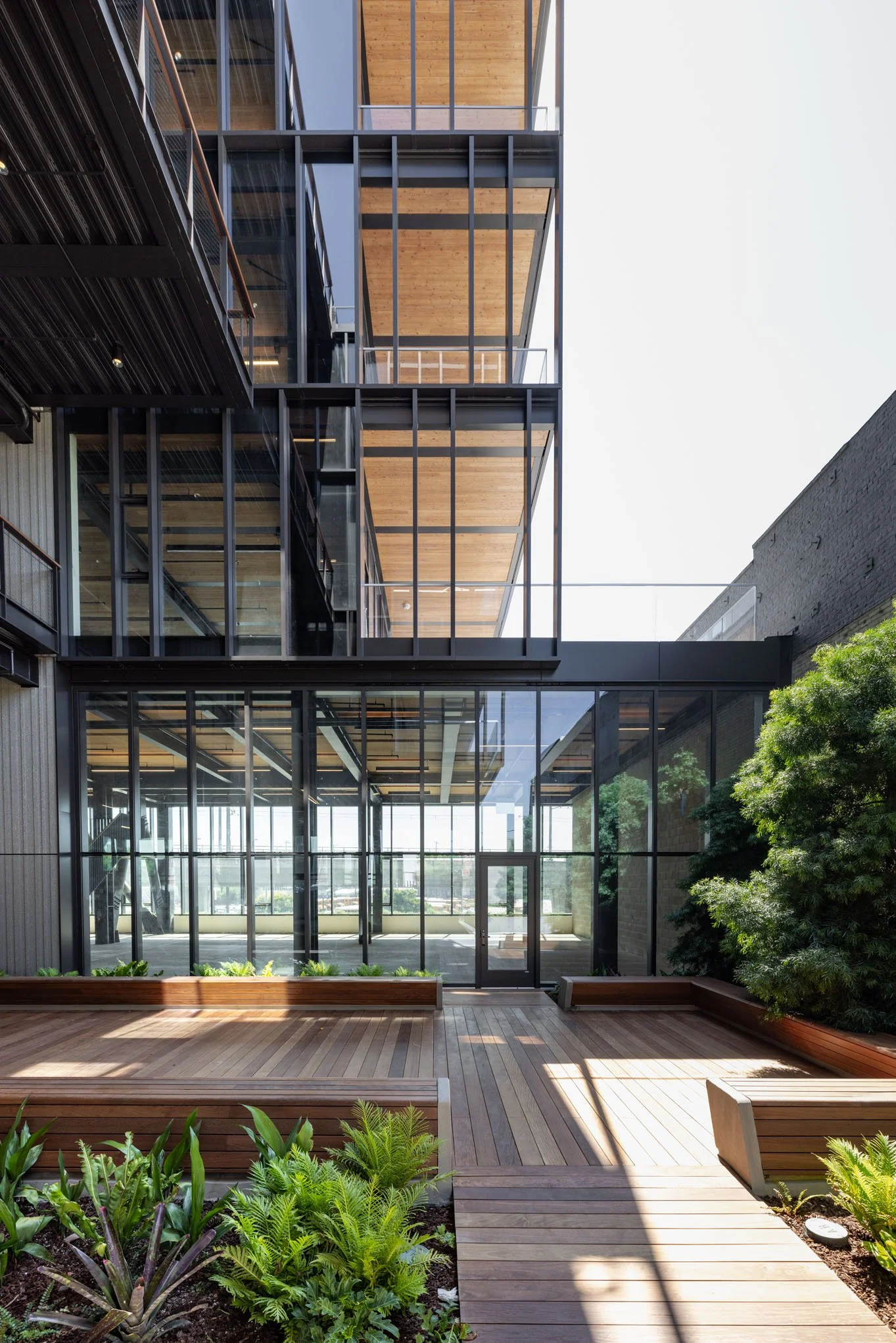 Modern multi-story building with glass exterior, wooden interior ceilings, and outdoor wooden deck with seating and green plants.