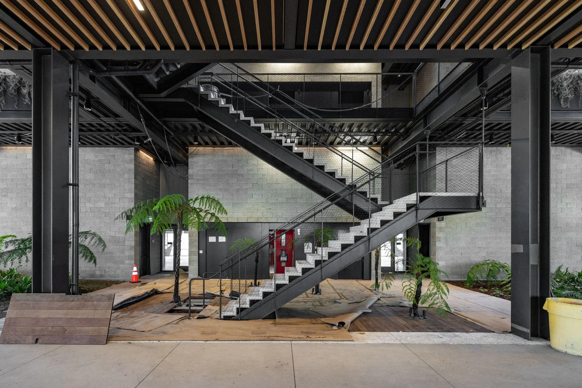 Interior of a modern building under construction with a black metal staircase, concrete floor, unfinished walls, plants, and construction materials.