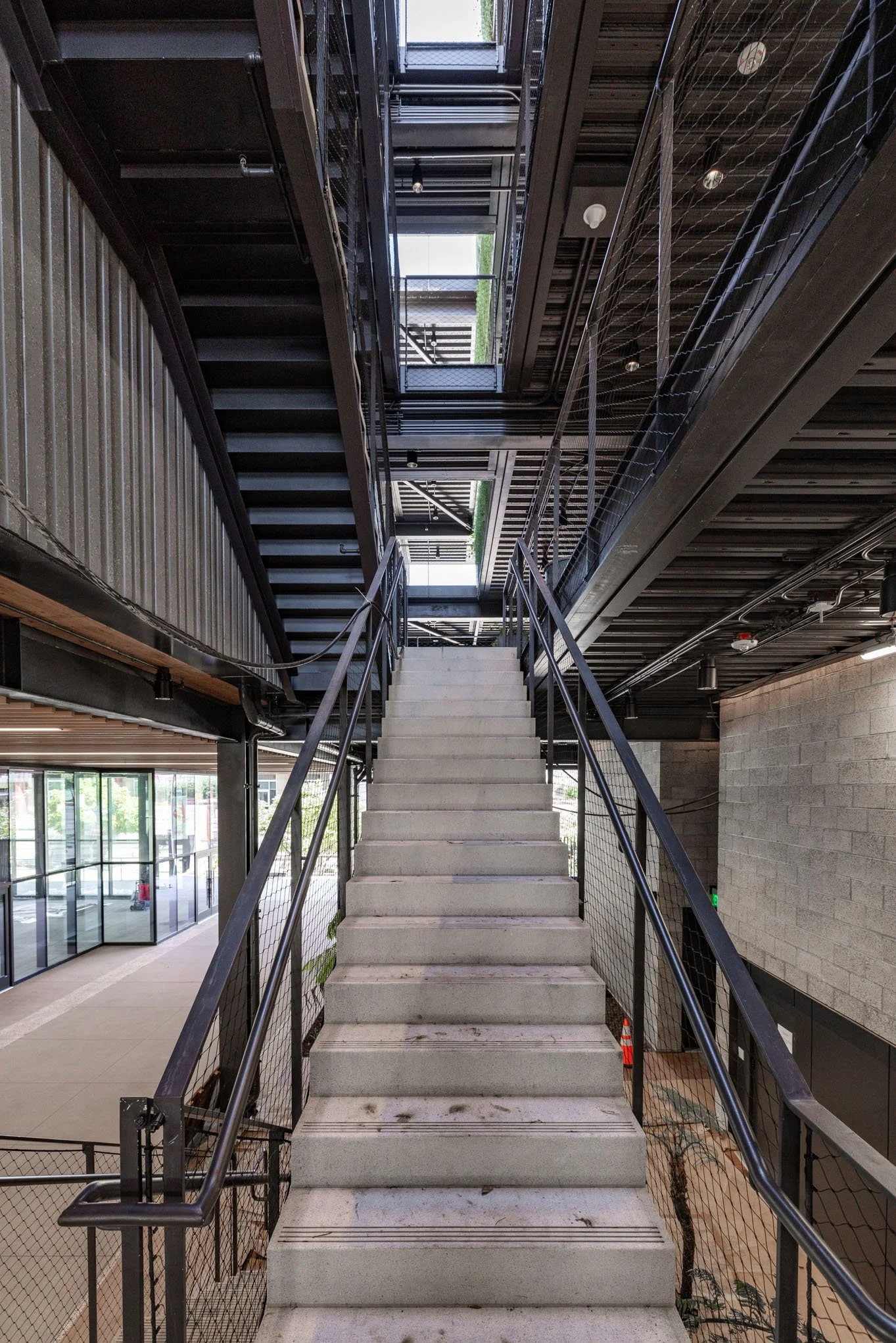 Indoor staircase with metal railings and open landings, leading upstairs in a modern building with exposed ceilings and large windows.