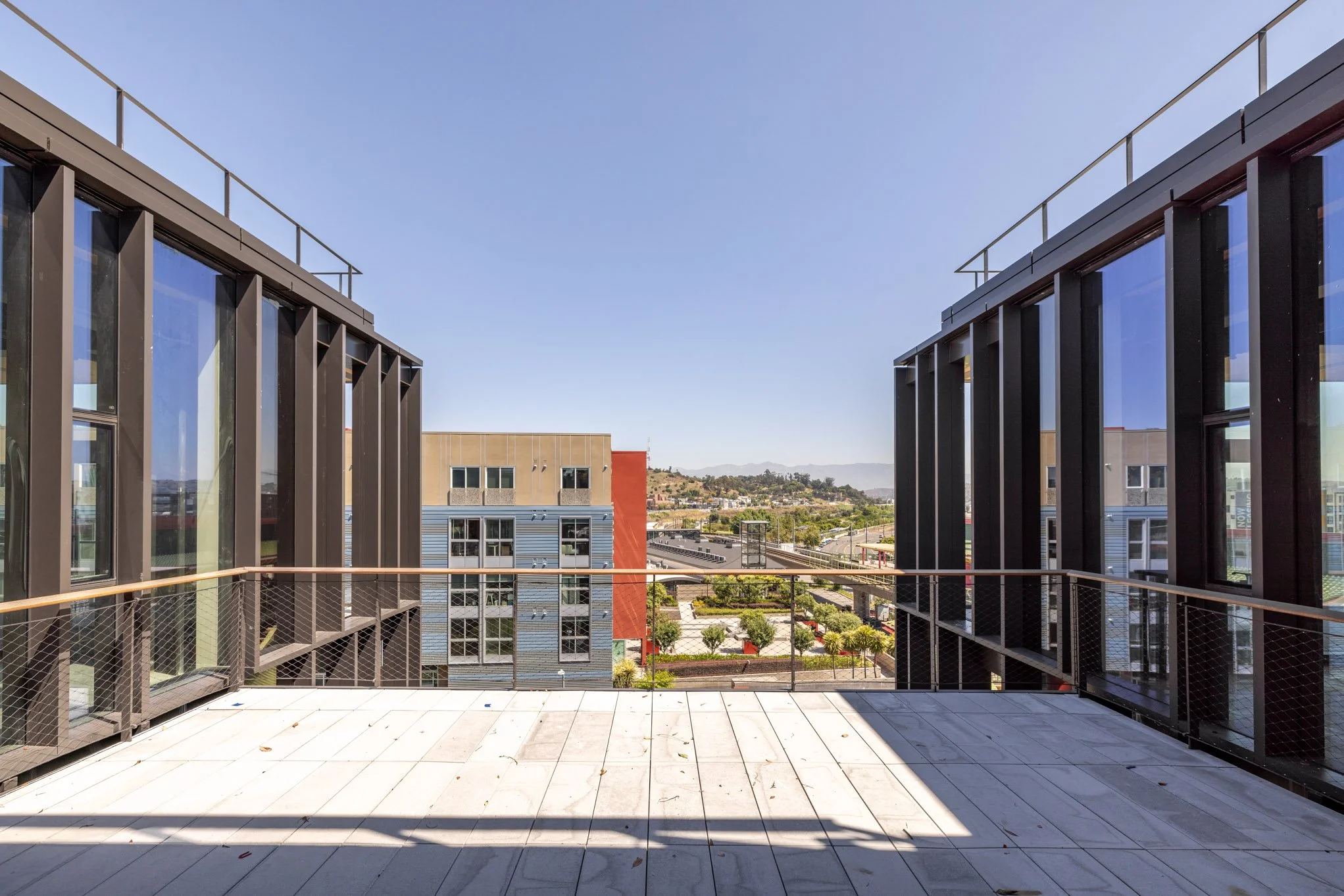 View from a balcony overlooking an urban area with modern buildings, trees, and distant hills on a clear, sunny day.