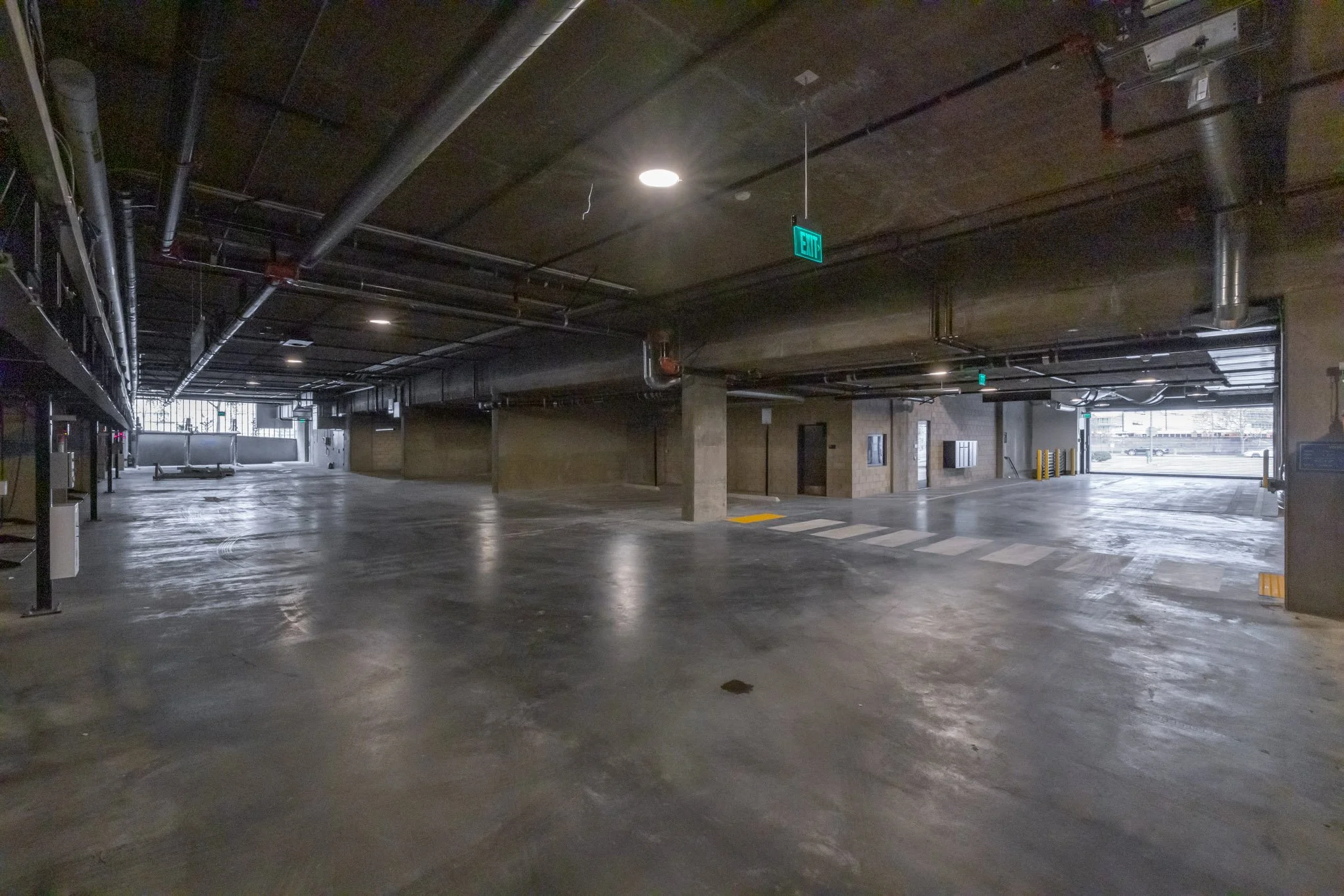 Empty indoor parking garage with concrete floors, exposed pipes on the ceiling, and an illuminated exit sign.