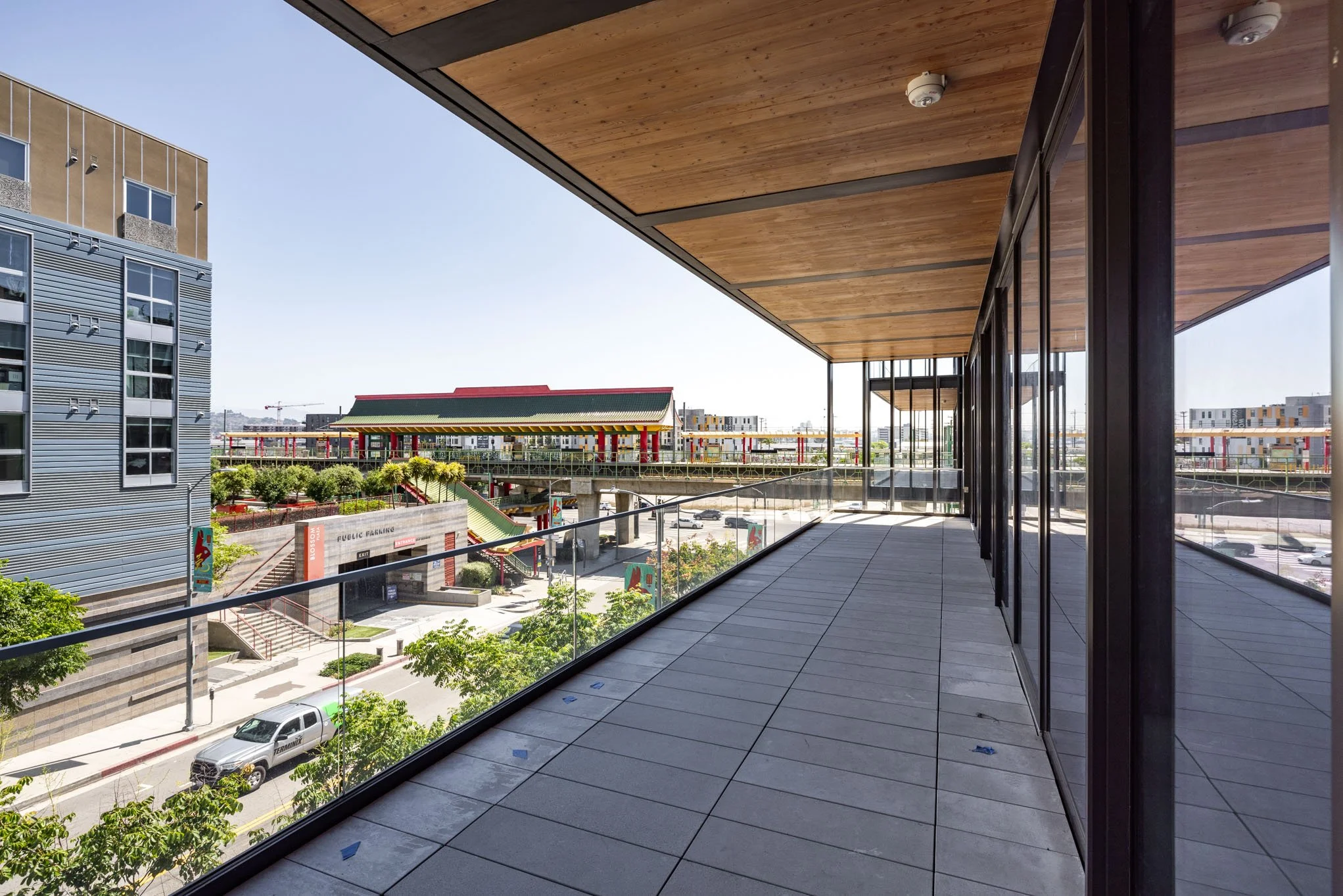 Empty balcony with glass railing overlooking an urban area with modern buildings, trees, and a parking lot.
