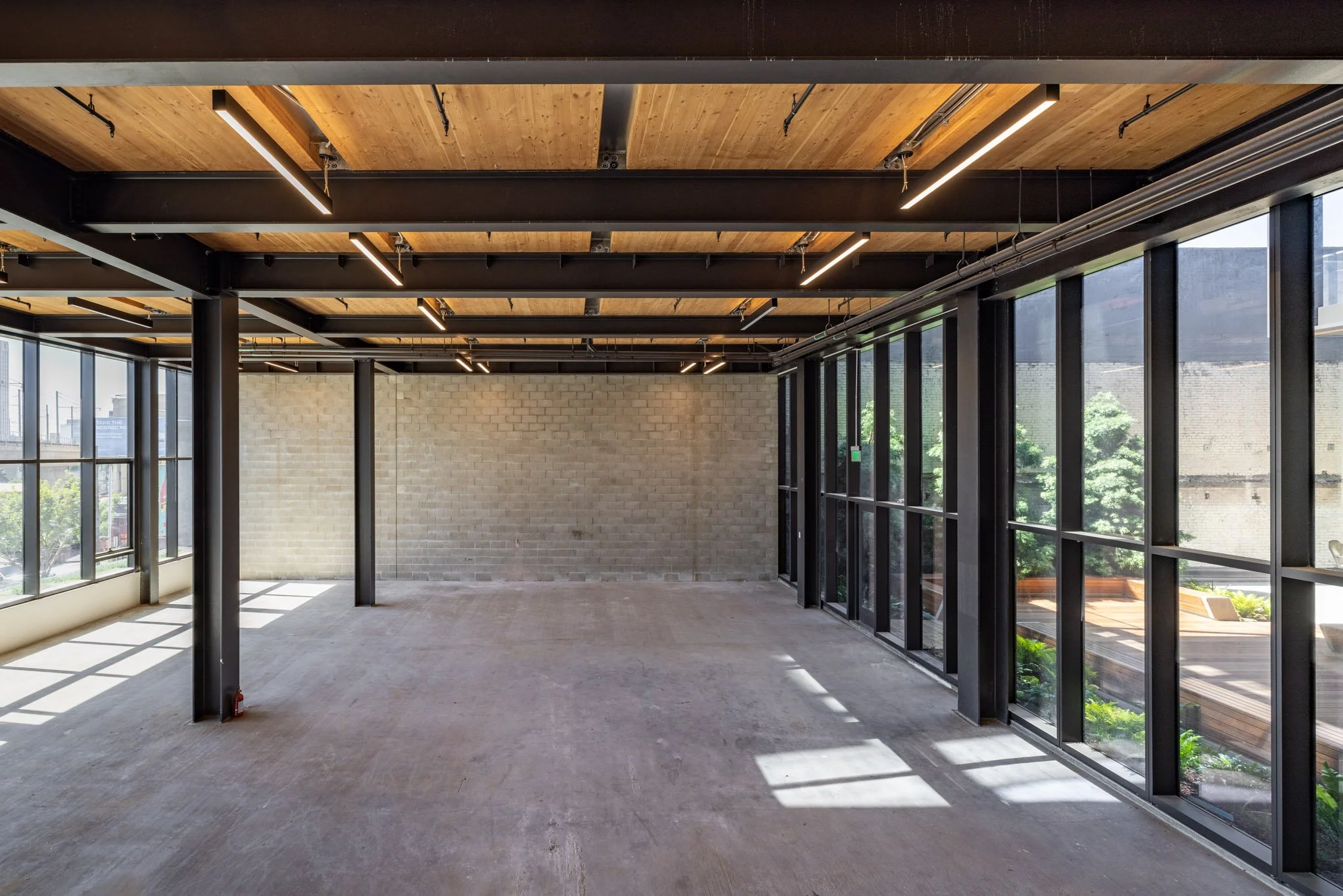 Empty commercial space with large windows, exposed ceiling beams, and unfinished floors.