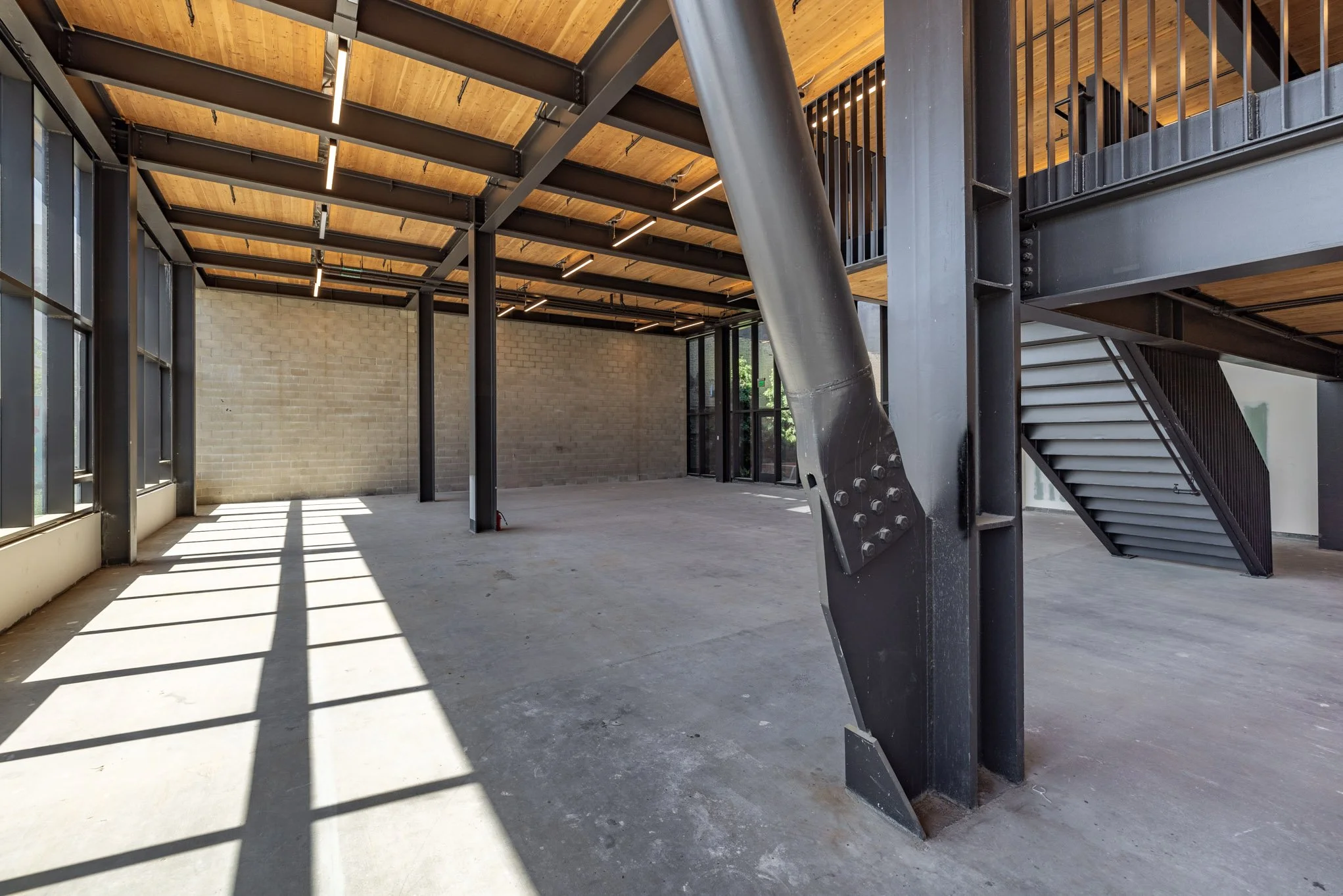 Empty interior space with concrete floor, brick wall, large windows, exposed steel beams, staircase, and natural lighting.