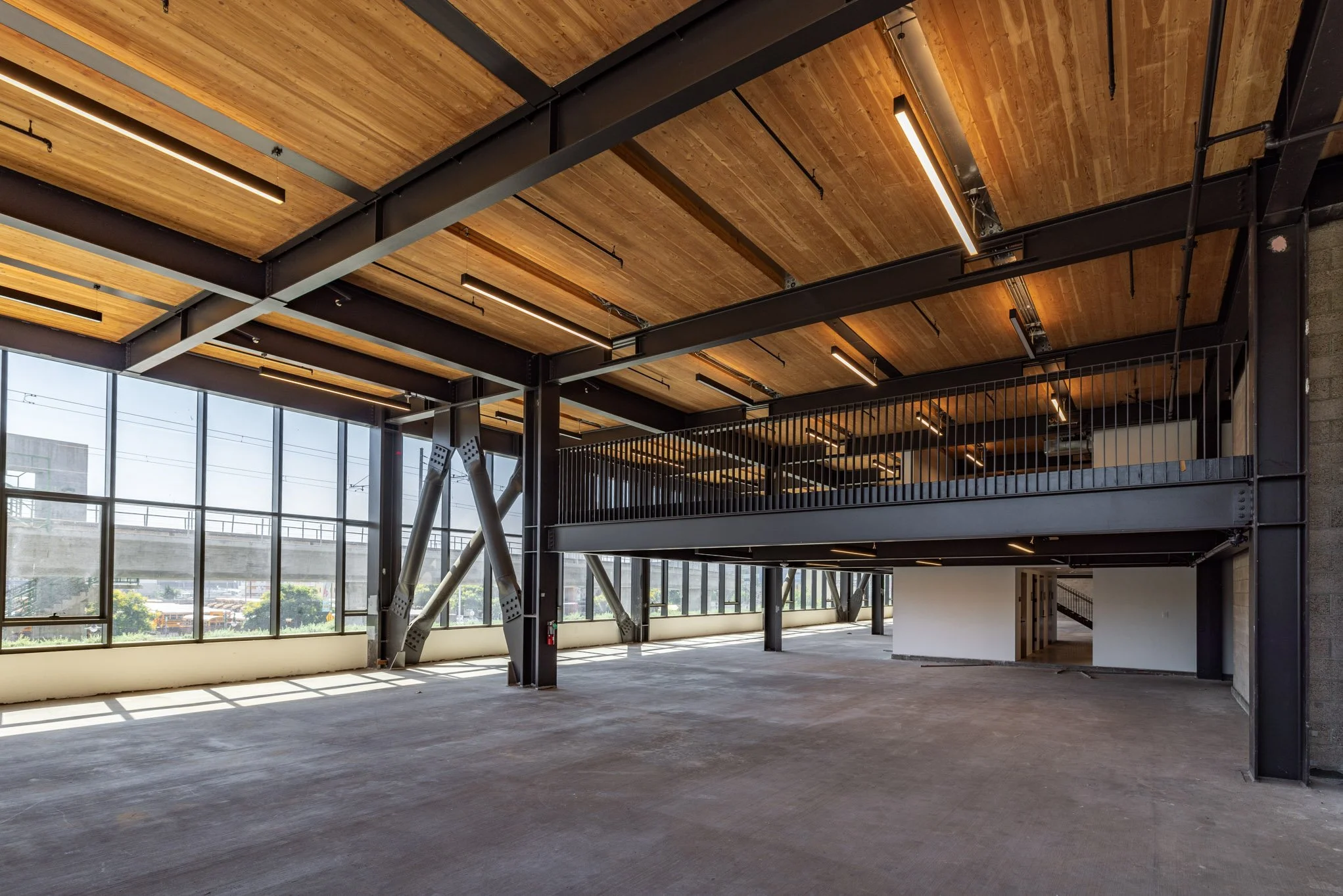 Interior view of an unfinished modern building with large windows, exposed metal beams, wooden ceiling, and a mezzanine level with black railing.