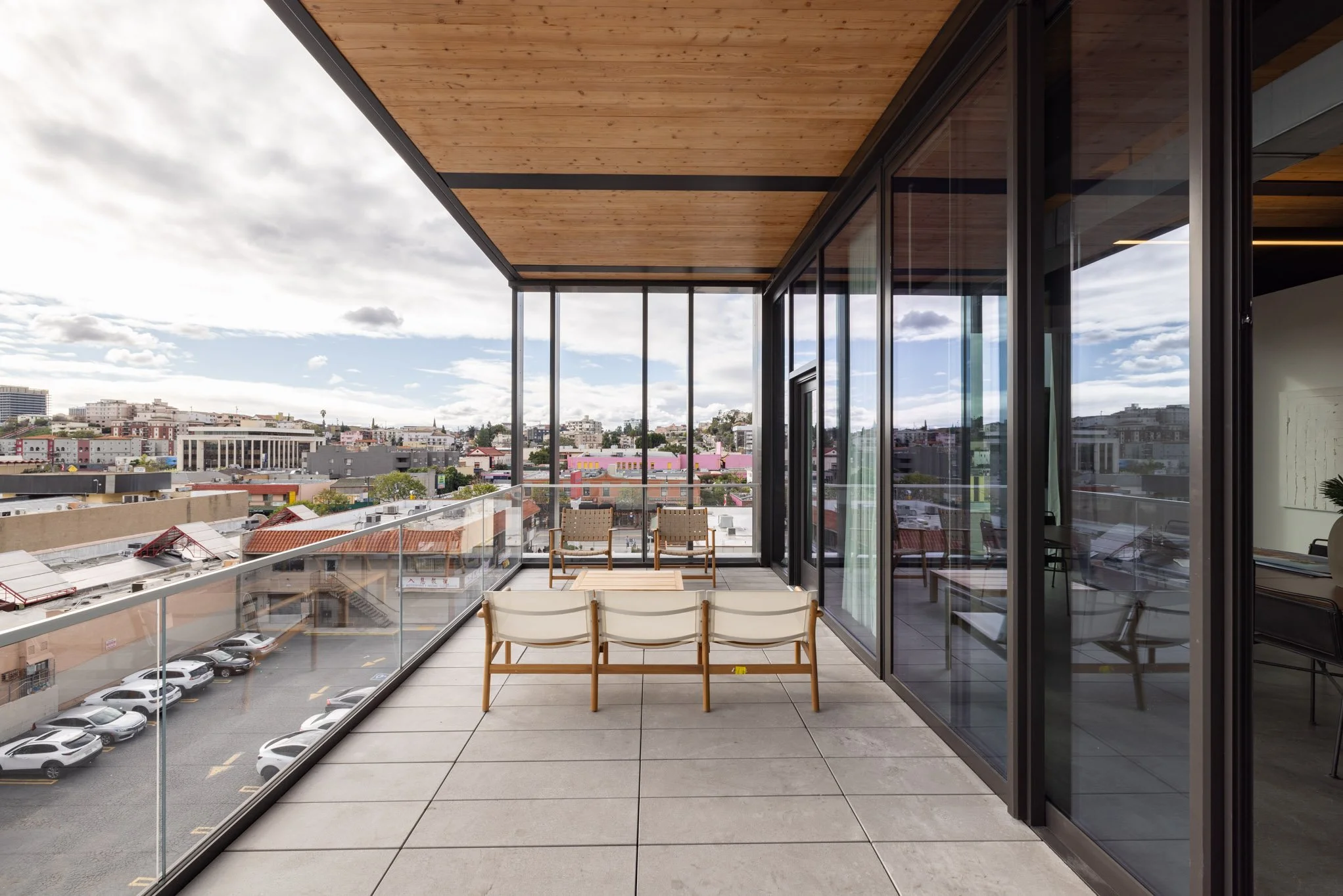 Empty balcony with outdoor seating, glass railing, and city view under cloudy sky.