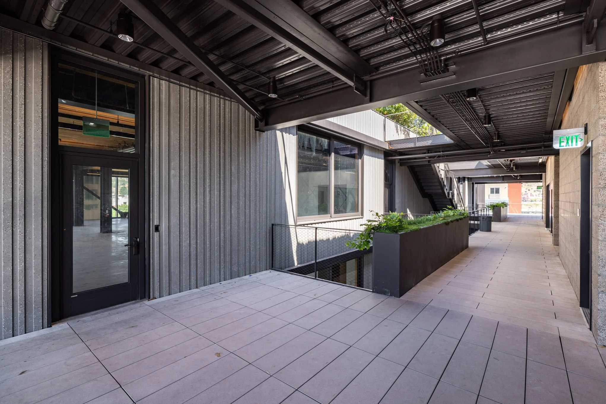 Modern outdoor corridor with metal siding, large windows, planter boxes with greenery, and exit signs.