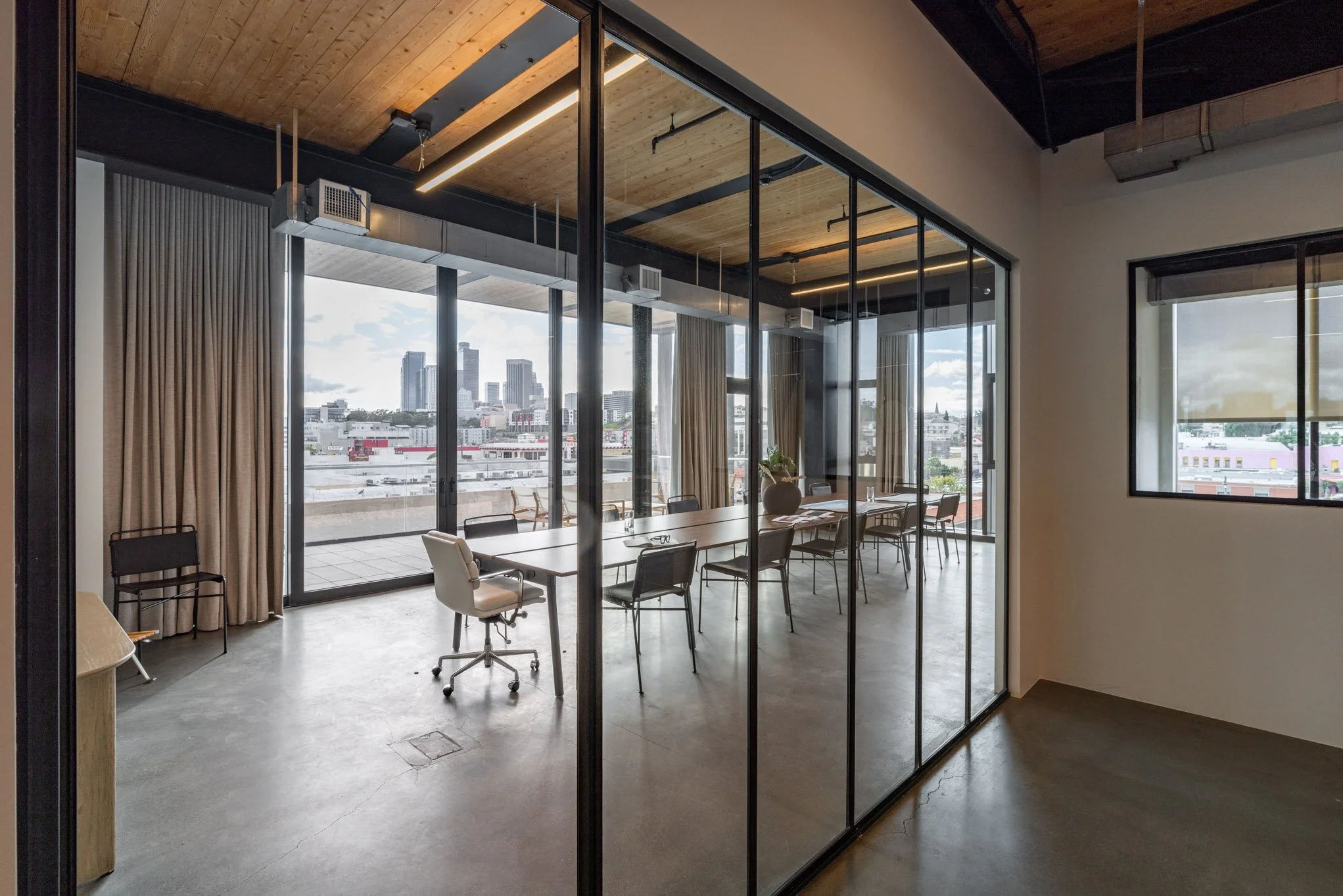 Modern office meeting room with glass walls, large window overlooking city skyline, long table with chairs, wooden ceiling, and concrete floor.