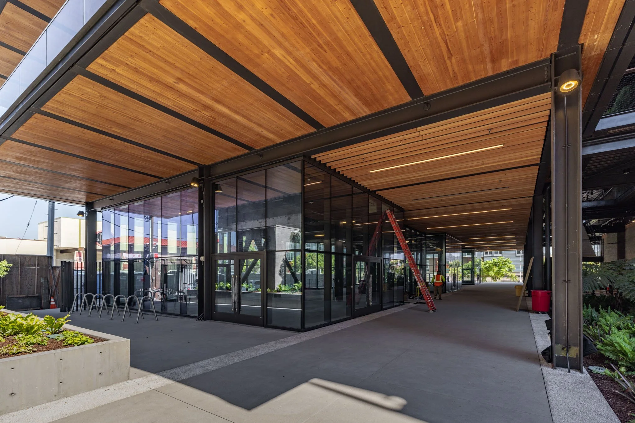 Construction site of a modern building with a wooden ceiling, glass walls, and a worker in a safety vest and helmet, with equipment such as a ladder and trash bin visible.