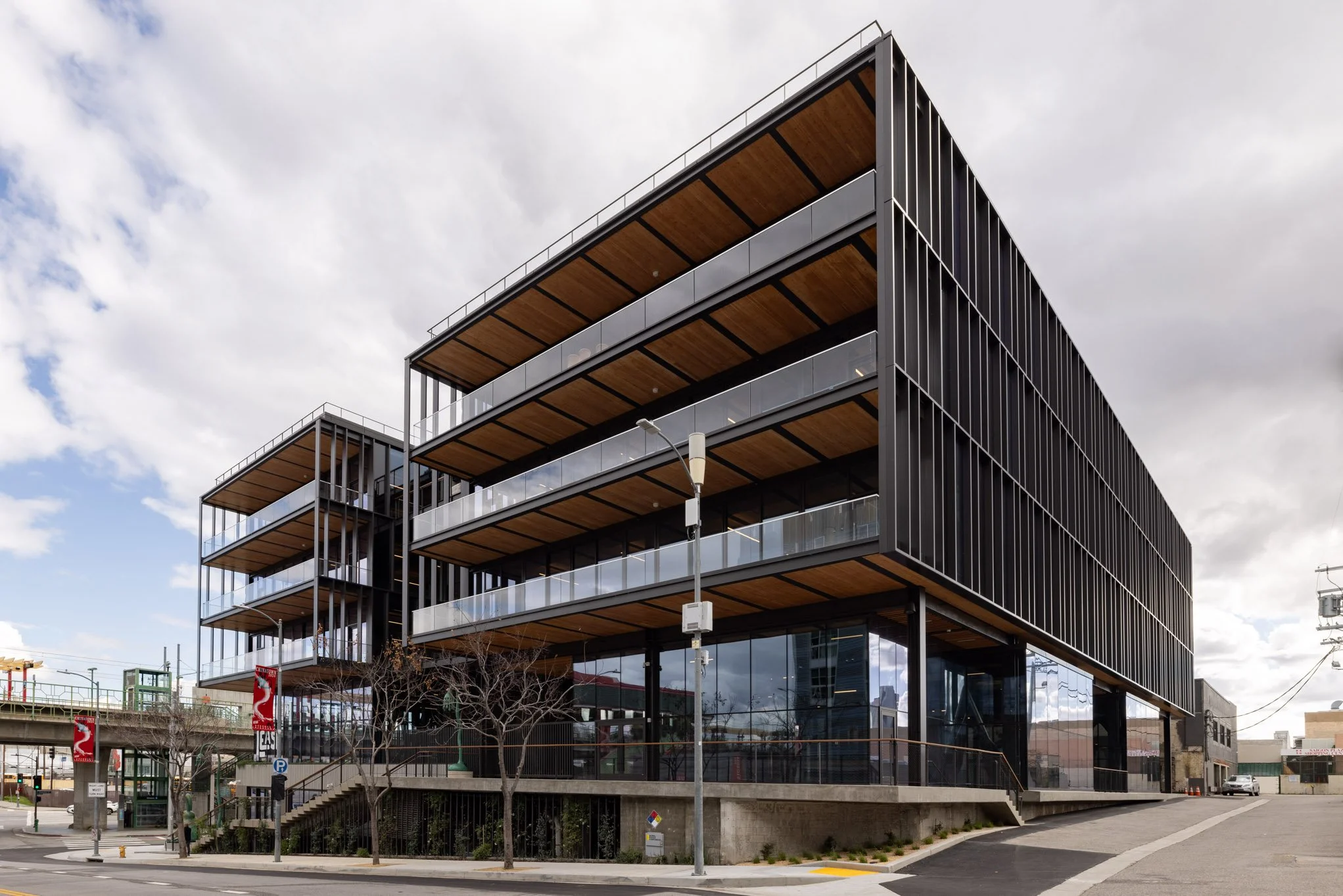 Modern multi-story building with black metal and glass exterior, large balconies, and wooden accents, situated on a city street with a few leafless trees and street lamps.
