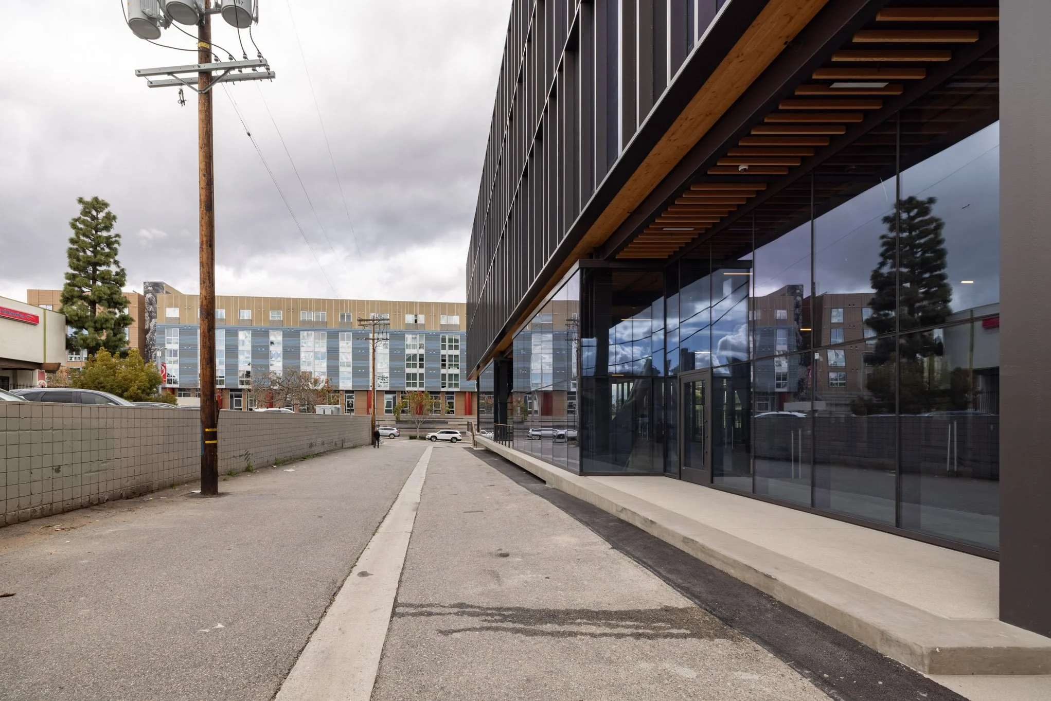 A modern building with large glass windows reflecting the cloudy sky, a sidewalk, utility poles, and a parking lot with a few cars.
