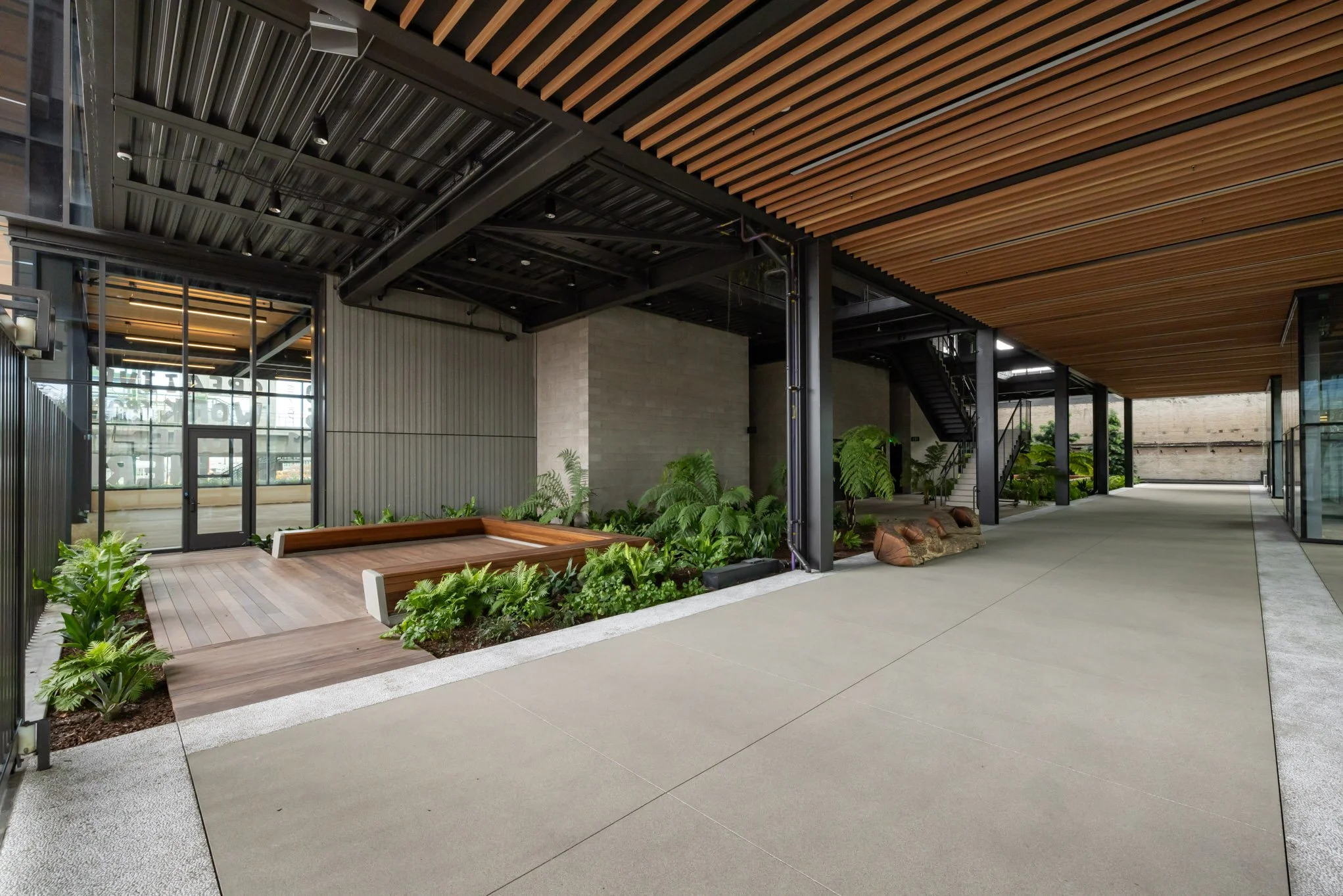 Modern outdoor area with a wooden bench, lush green plants, and a covered patio with metal and wood accents.