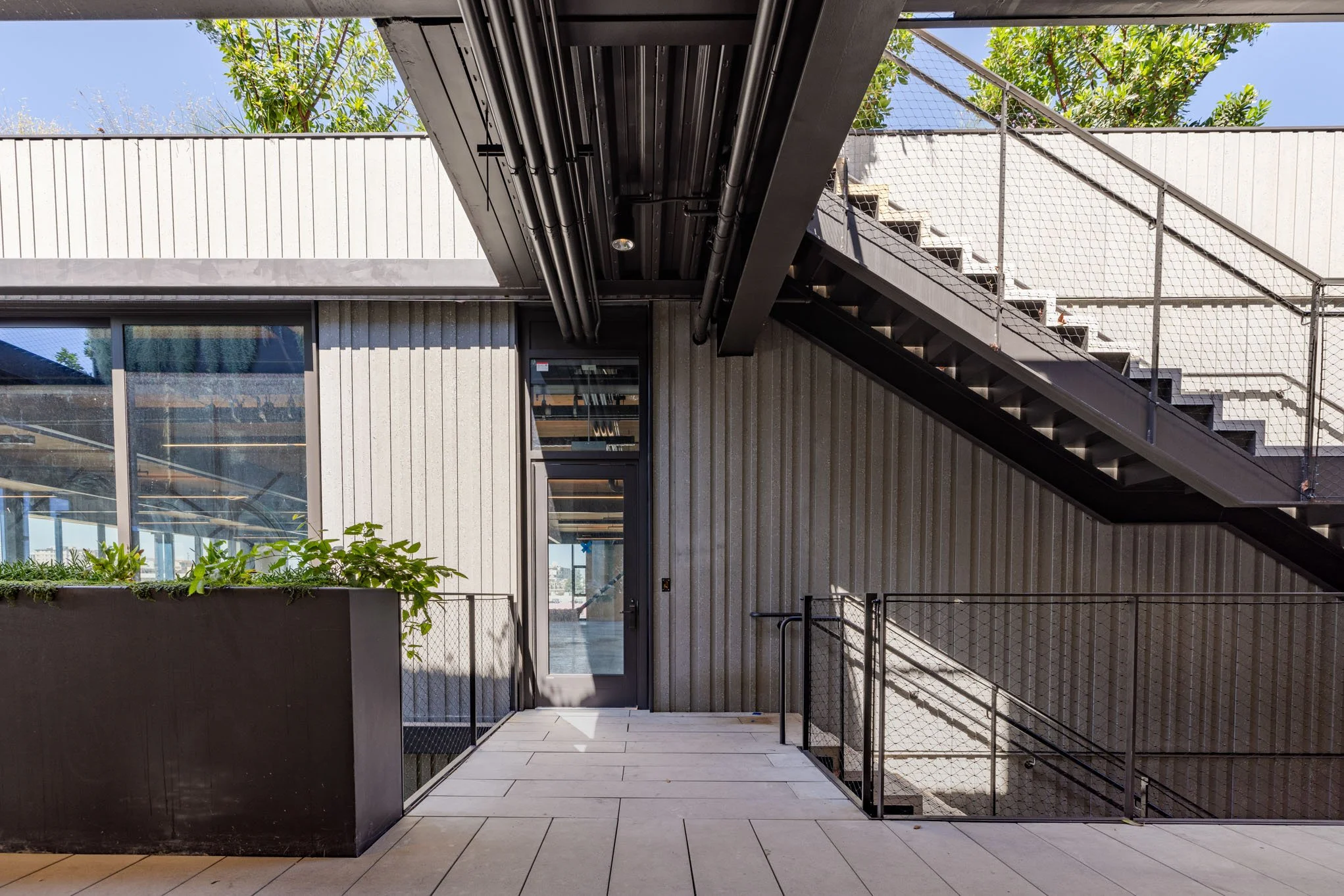 Modern building exterior with outdoor staircase, black railings, large window, and potted greenery, under a clear blue sky.