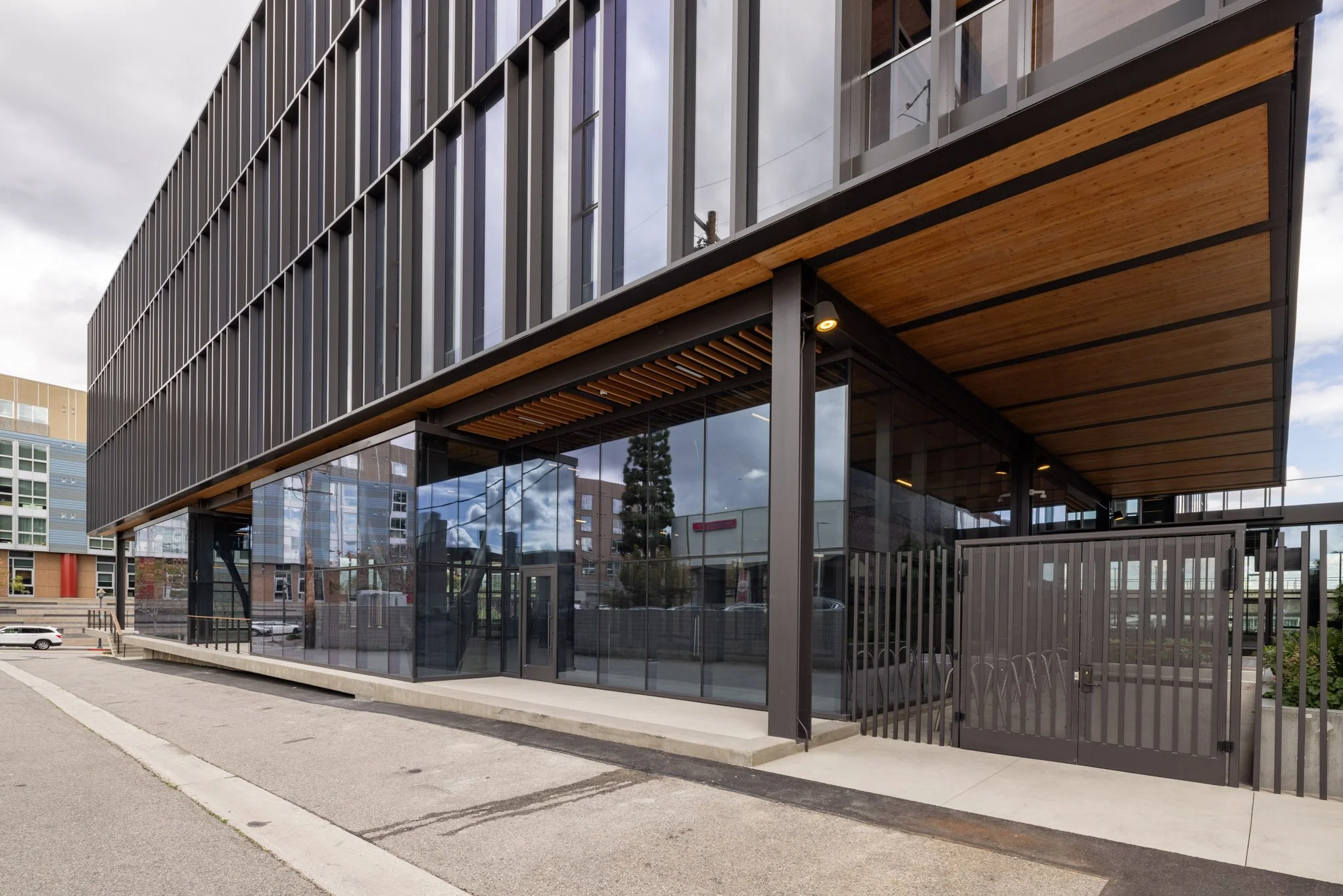 Modern multi-story building with large glass windows, wooden accents, and a gated entrance, located in an urban area.