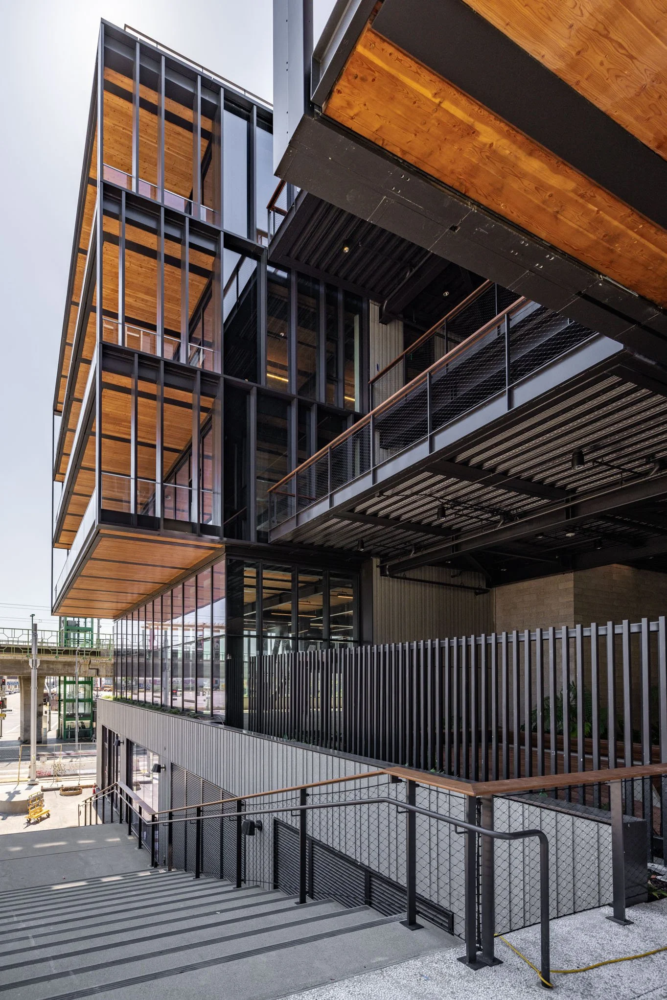 Modern multi-story building with glass and metal exterior, featuring wooden balcony accents and a stairway outside.