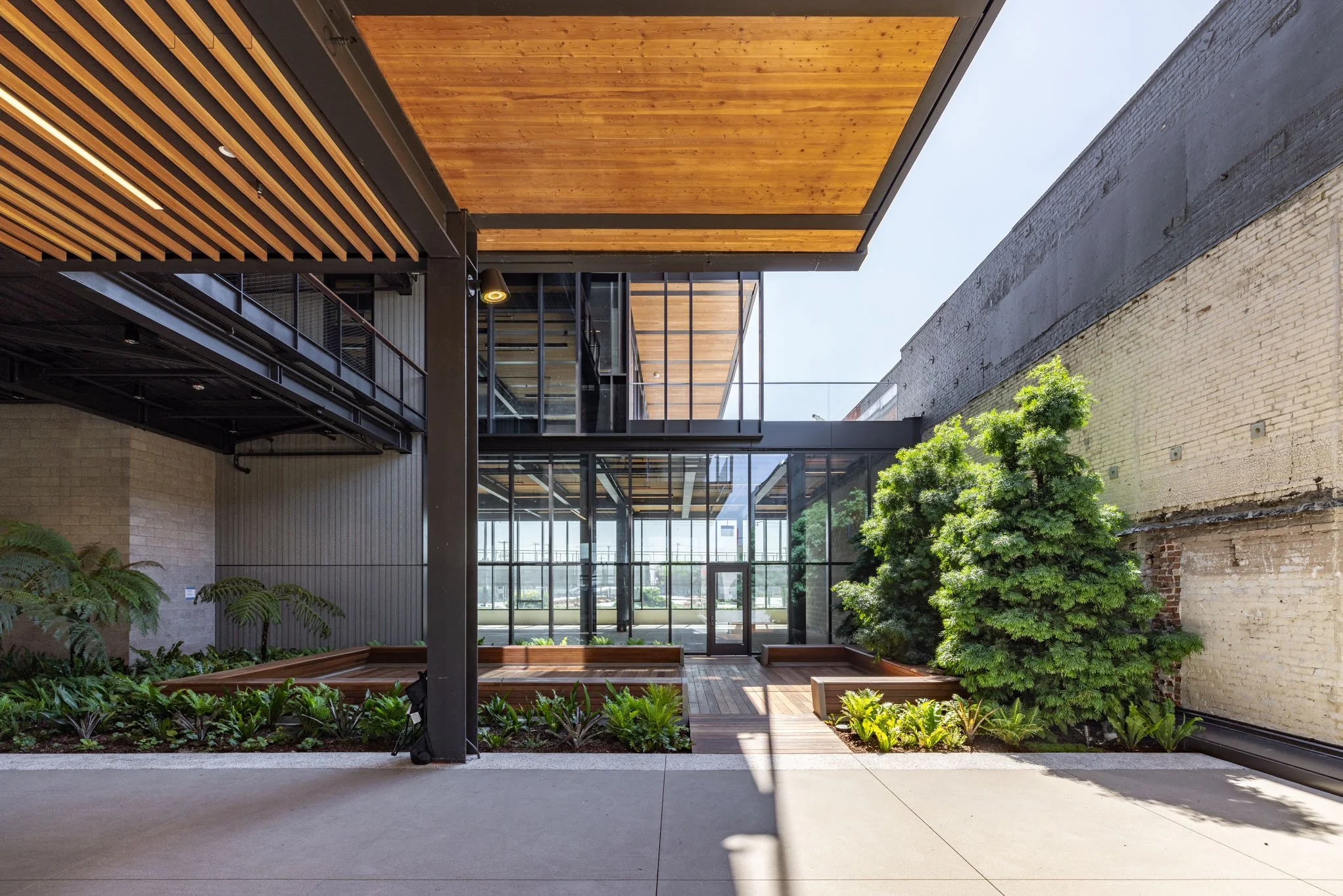 Modern architectural courtyard with glass walls, wooden ceiling panels, green plants, and brick walls.