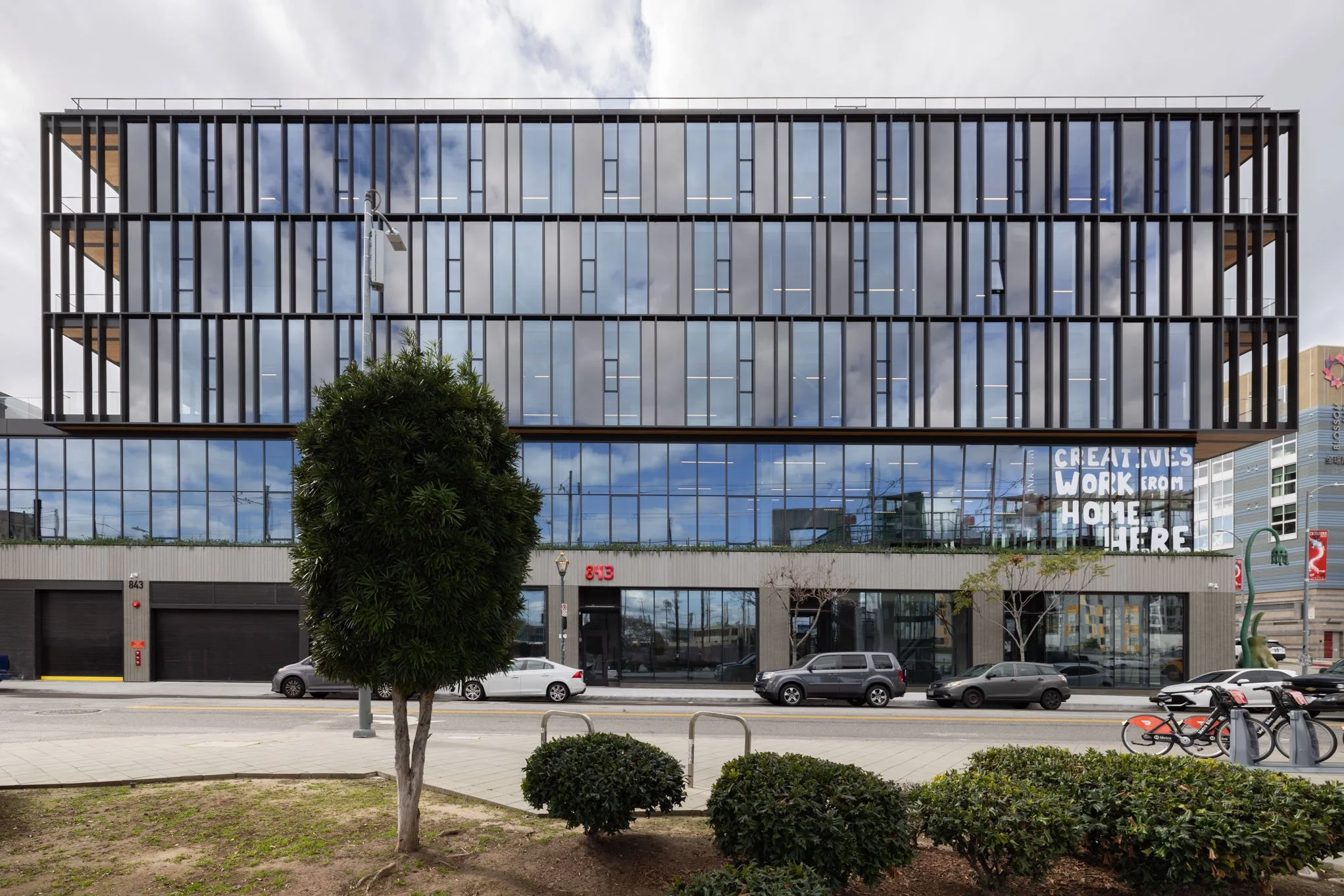 Modern multi-story glass office building with a sign in the window reading 'GREAT JOBS WORK FROM HOME HERE!', parked cars, trees, and bicycles in front.
