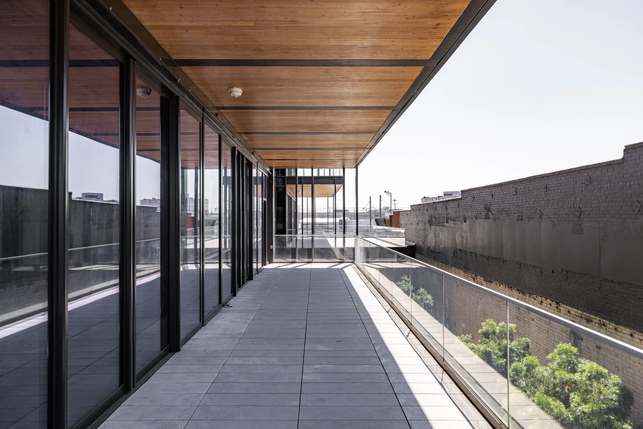 Modern balcony with glass railing, tiled floor, and wood ceiling, overlooking cityscape with buildings and trees.