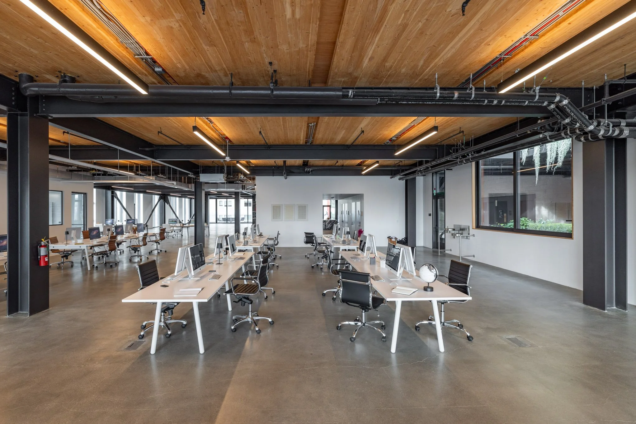 An open-concept modern office space with white desks, black swivel chairs, and desktop computers. The ceiling is wood-paneled with black industrial pipes and lighting. Large windows allow natural light, and a fire extinguisher is mounted on a black c
