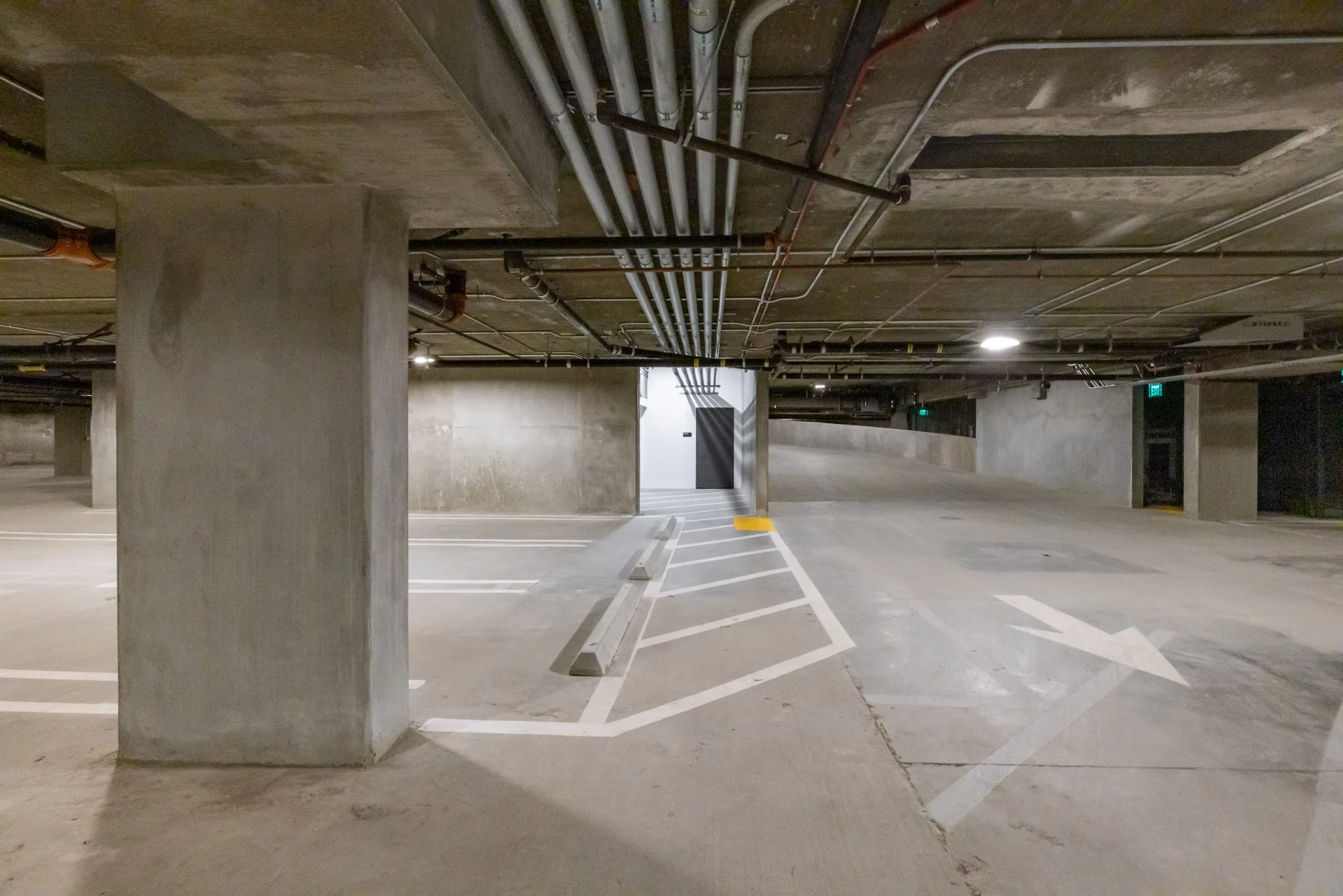 Empty underground parking garage with concrete pillars, painted parking lines, and a ramp leading upward, illuminated by ceiling lights.