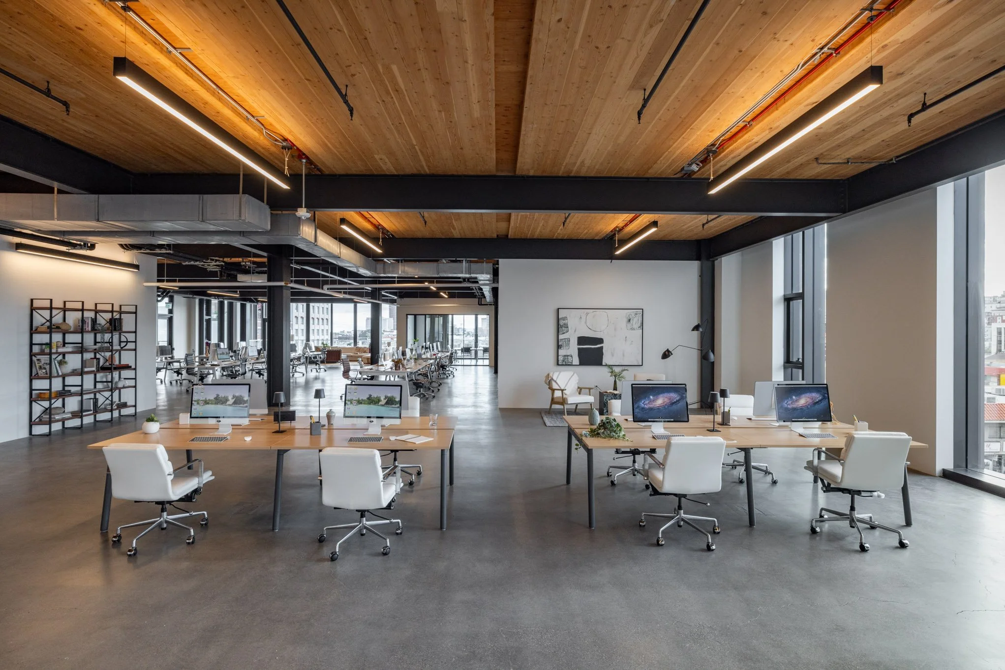 Modern open office space with desks, white swivel chairs, computer monitors, large windows, and minimalistic decor.