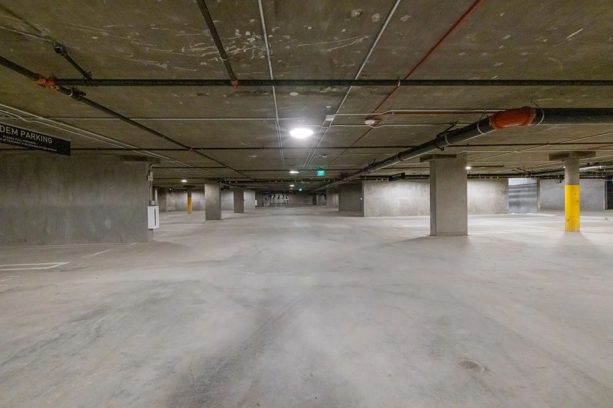 Empty underground parking garage with concrete pillars and ceiling pipes, dim lighting, and a green exit sign.