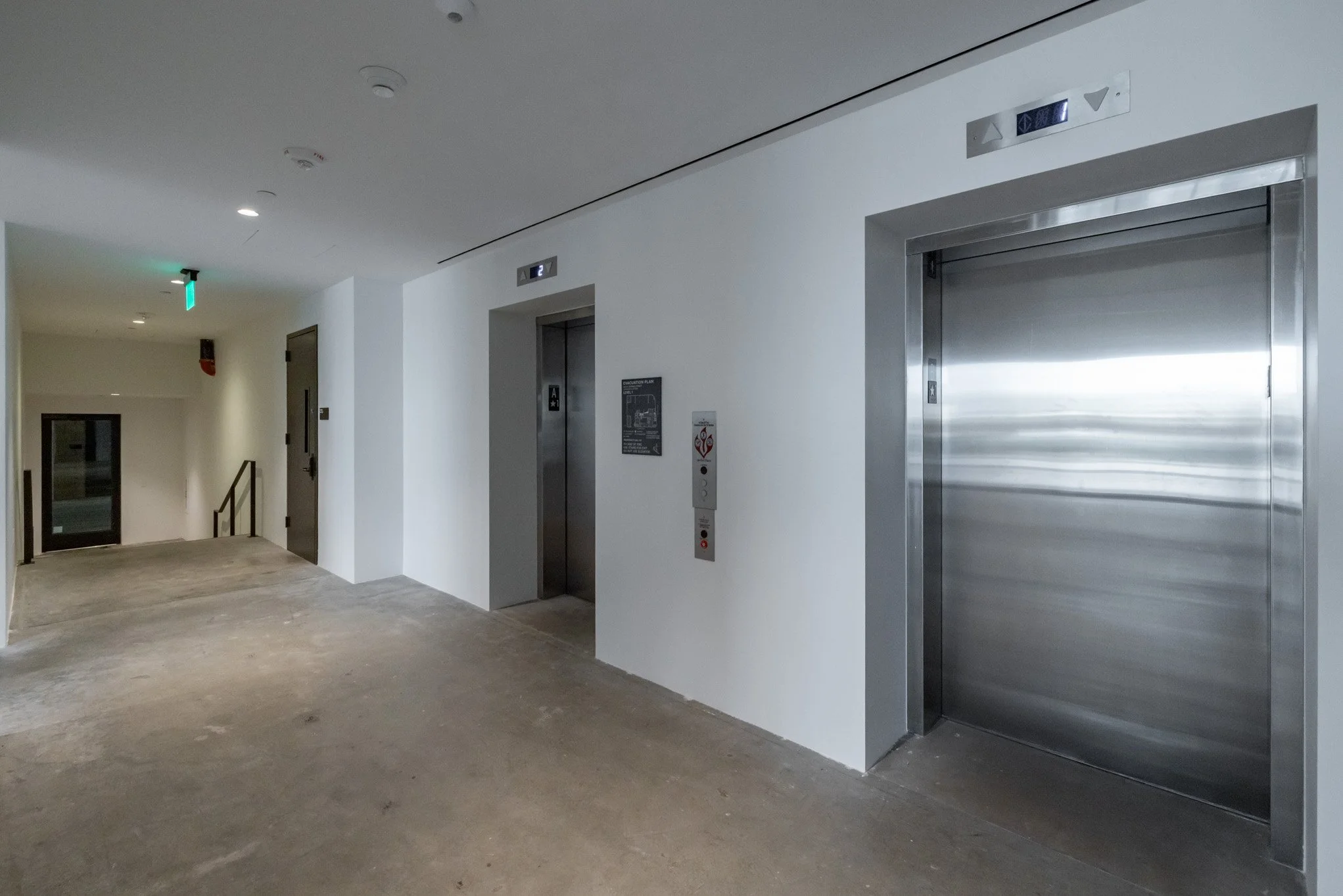 Interior of a building with two elevator doors, an emergency exit door, and a staircase. The floor is unfinished concrete. The walls are white. The elevator on the right is closed, the one on the left is open showing a black interior with an elevator
