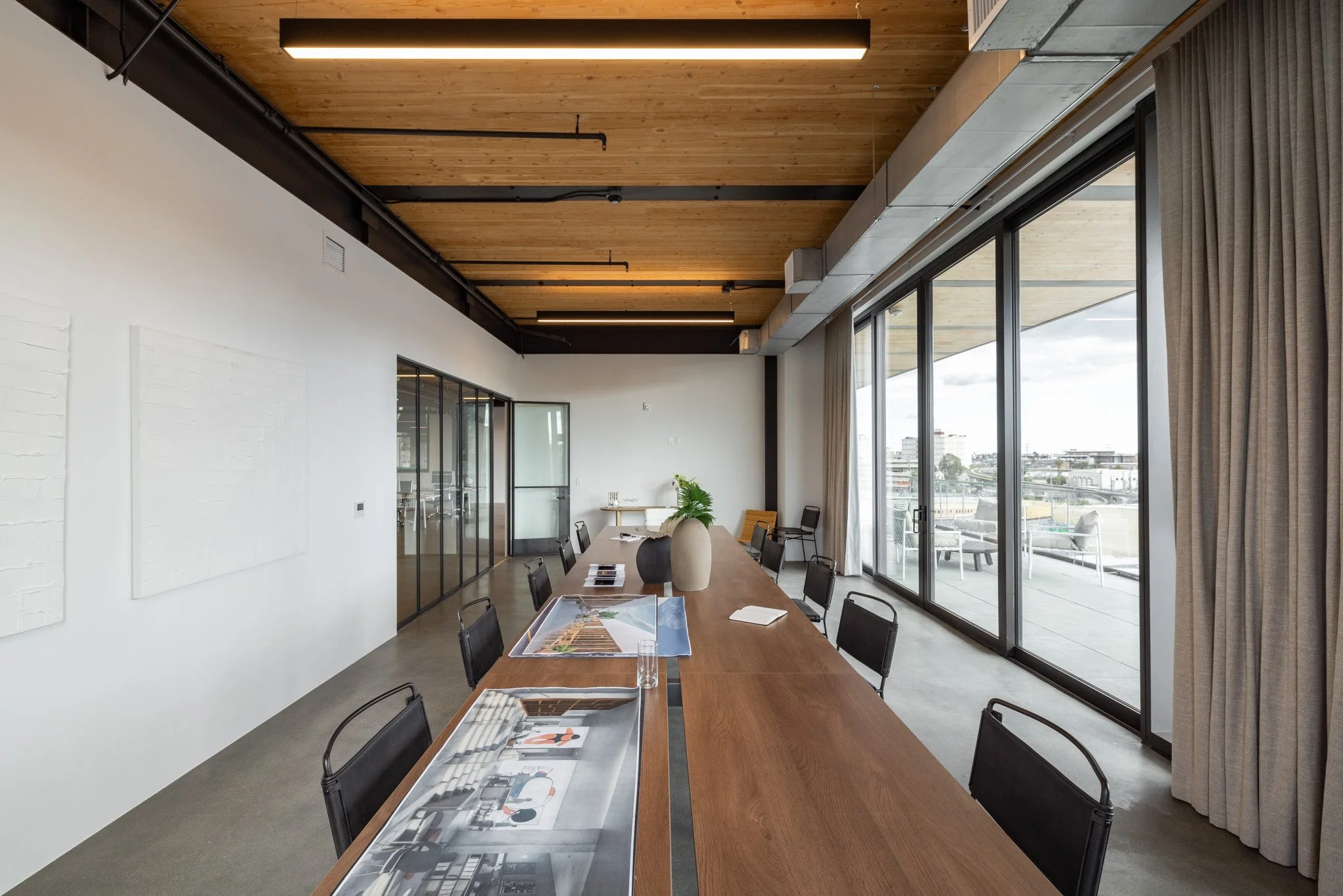 Empty conference room with wooden ceiling, large glass windows, and a long wooden table with black chairs, vases, and documents on it.