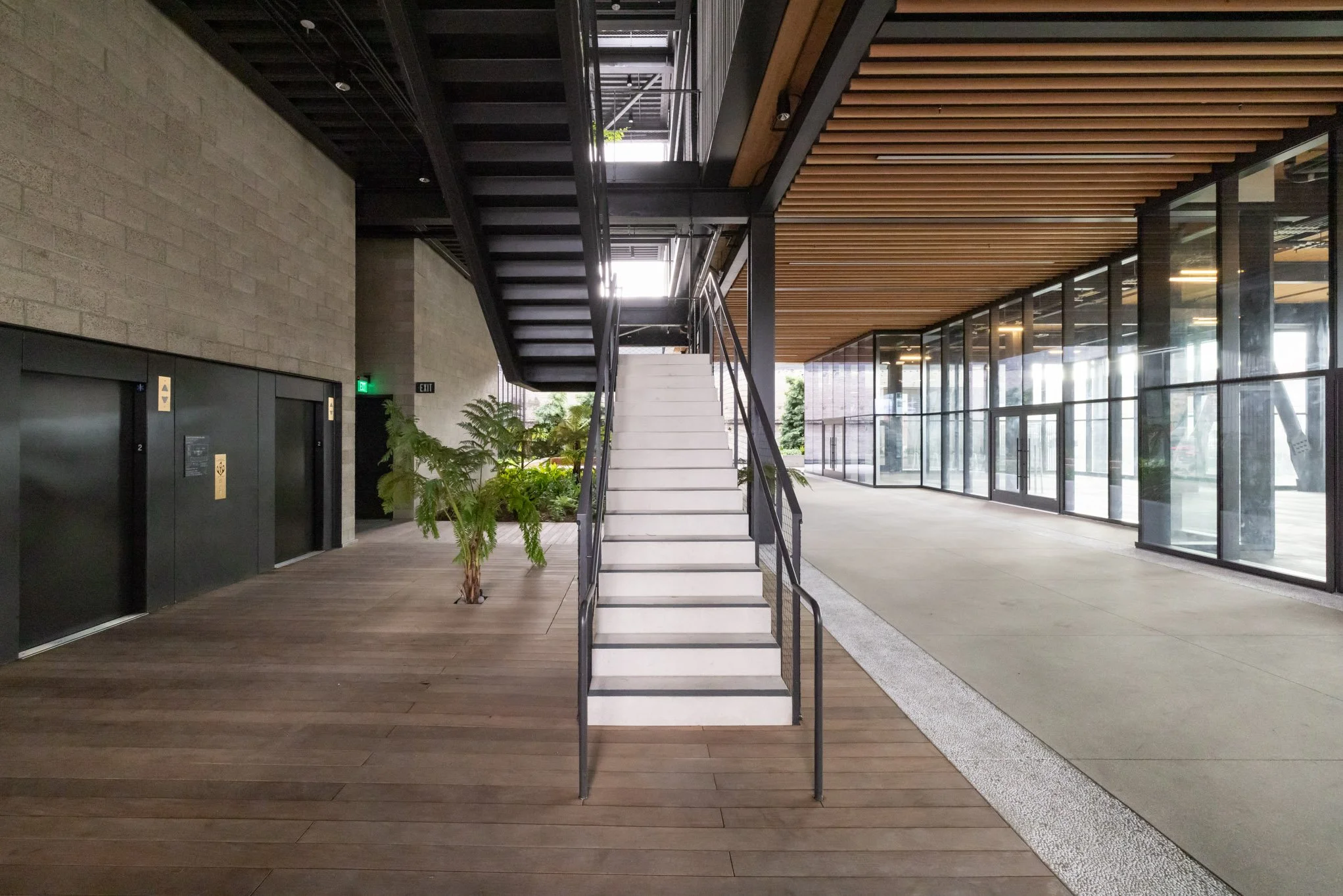Interior of a modern building lobby with a staircase, glass walls, plants, and elevators.