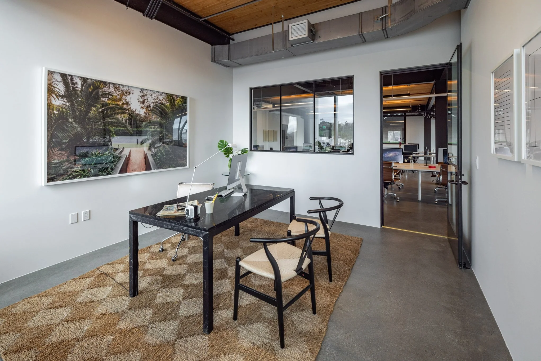 Modern office with a black desk, white chair, two wooden chairs, large landscape artwork, and a rug on a concrete floor.