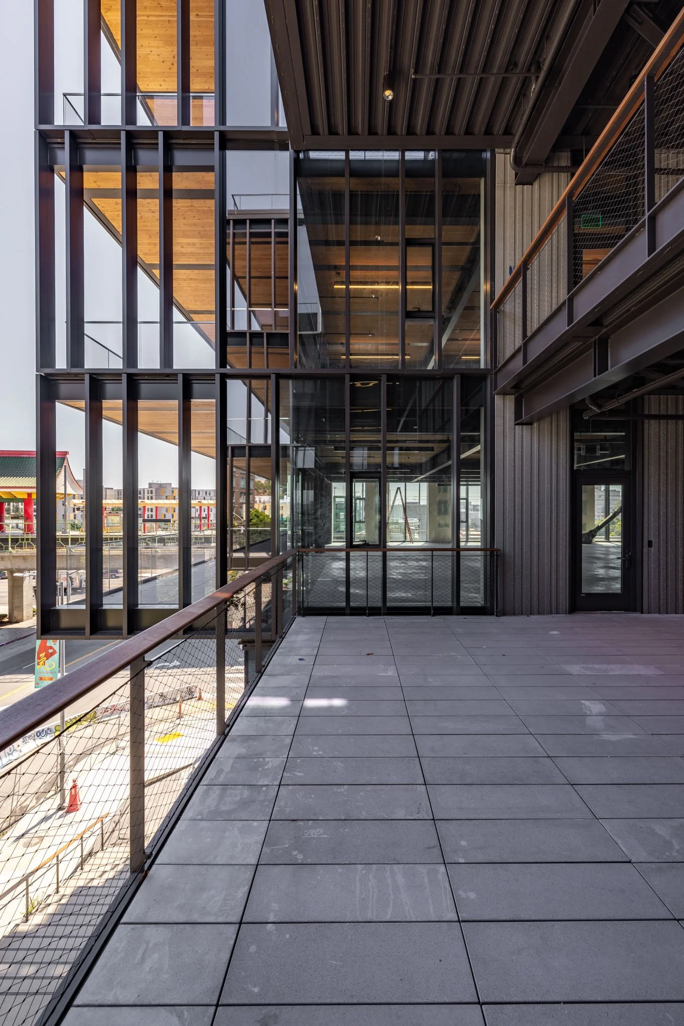 Modern building with glass windows, metal framework, and wooden accents, viewed from a balcony with tiled flooring and a safety railing.