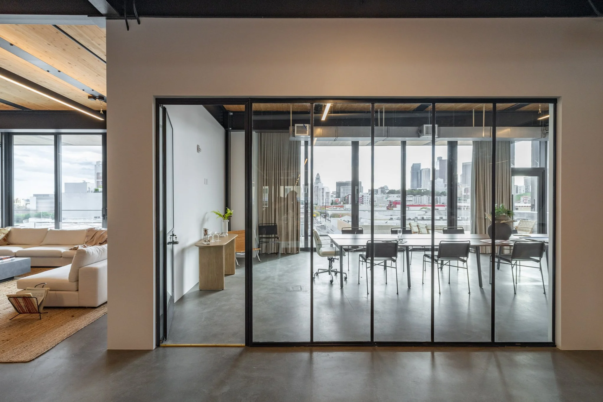 A modern conference room with glass walls, a long table, and chairs, with a city skyline view outside.