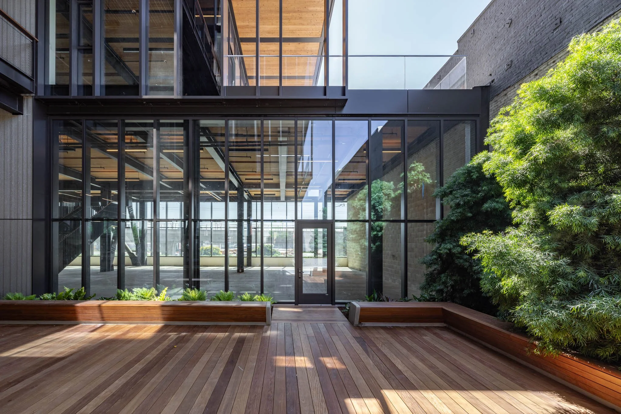 Modern building with floor-to-ceiling glass windows, wooden decking, and green plants outside.
