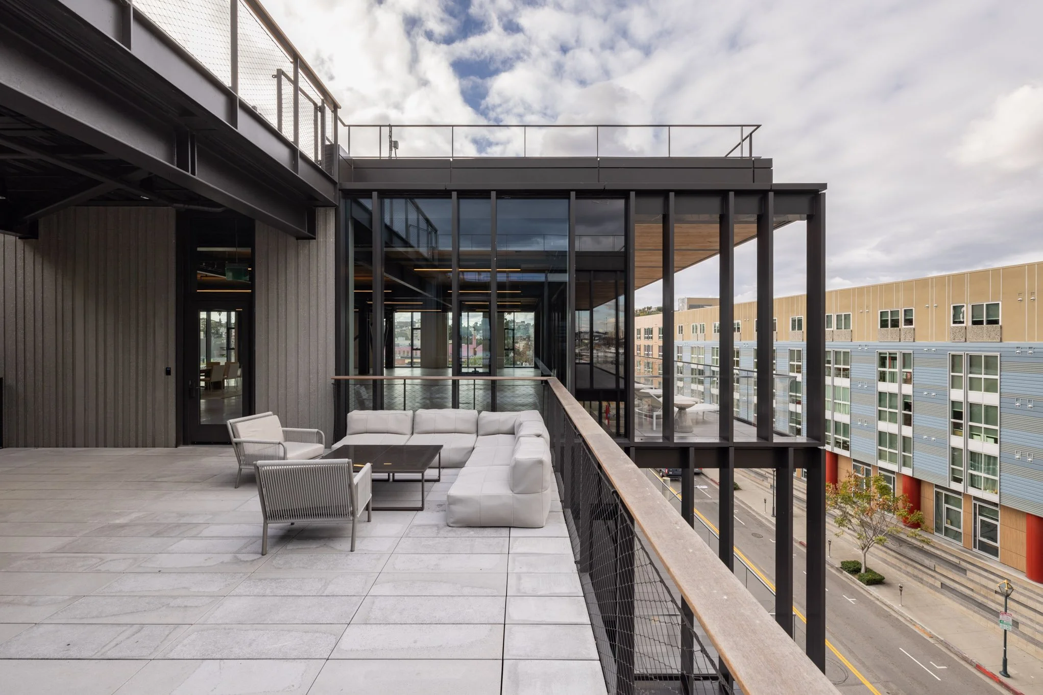 Outdoor balcony with modern white sectional sofa, chairs, and black coffee table, overlooking city street with contemporary buildings and cloudy sky.