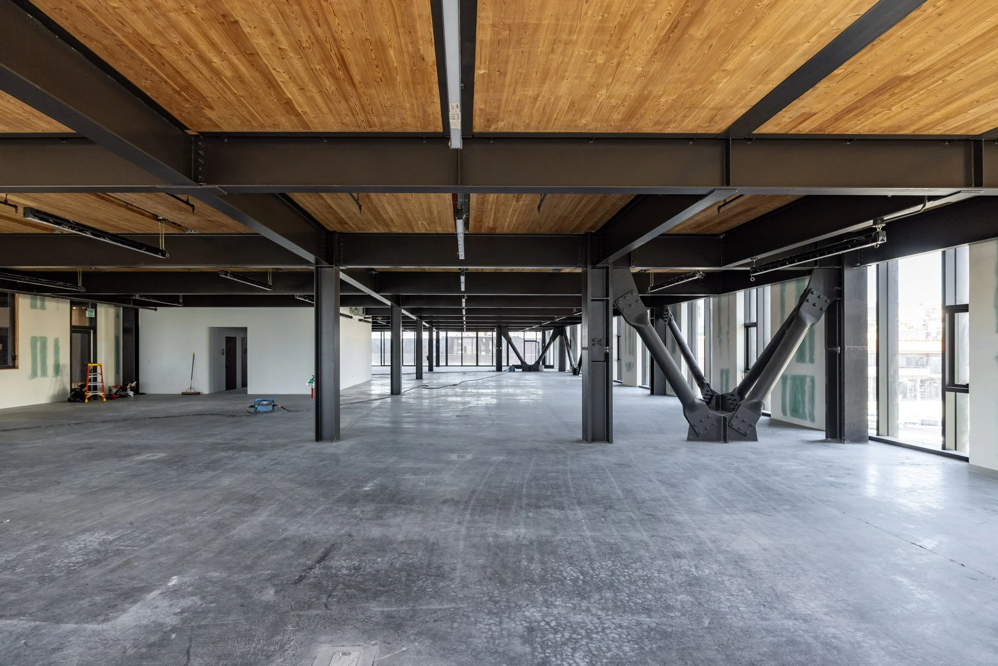 Interior view of a building under construction with exposed steel beams, wooden ceiling panels, concrete floor, large windows, and construction tools.