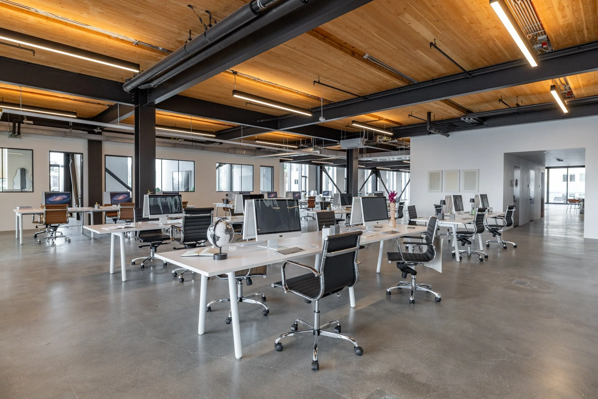 Modern open-plan office with white desks, black swivel chairs, multiple desktop computers, and large windows, featuring a wooden ceiling and industrial-style black metal beams.